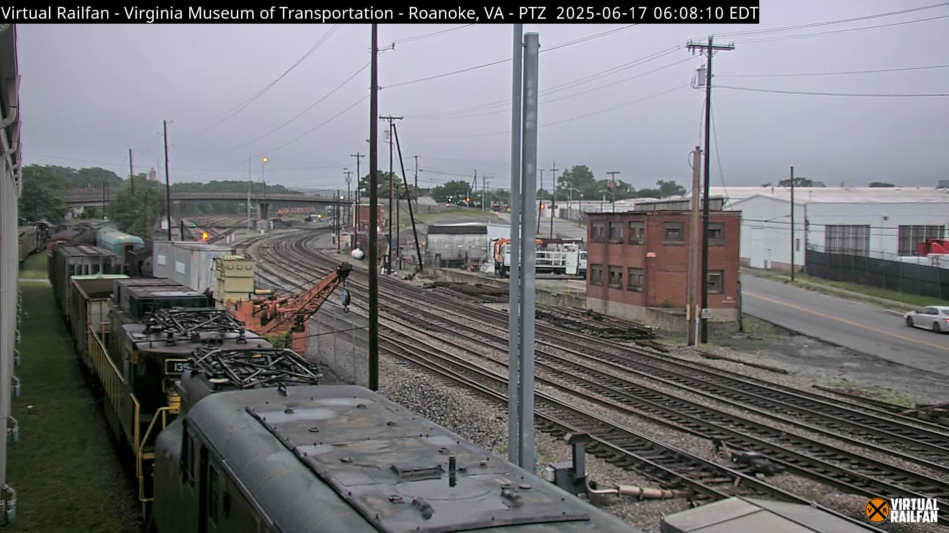 A long freight train sits on multiple railway tracks next to an overcast sky, with industrial buildings and a road visible in the background.