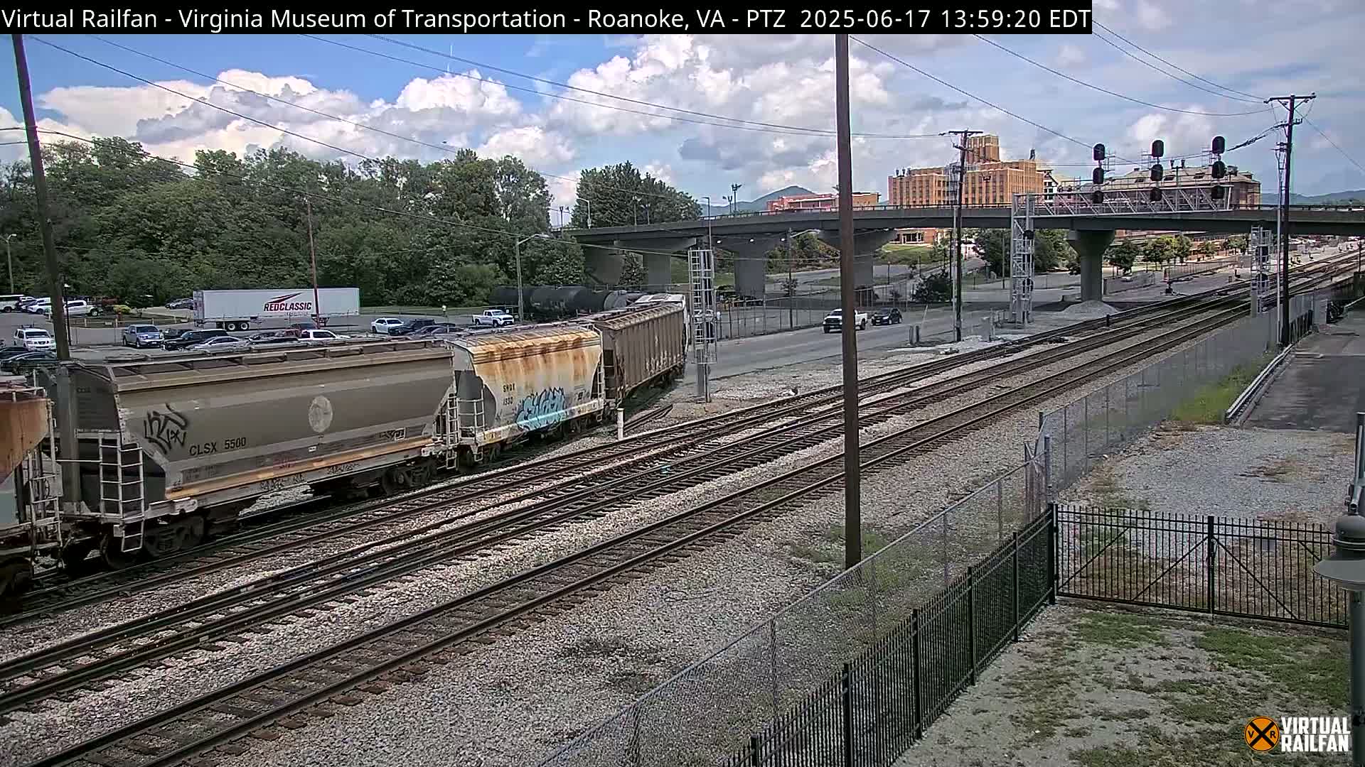 Several freight train cars sit on railway tracks under a partly cloudy sky, near a highway overpass and a parking lot with cars.