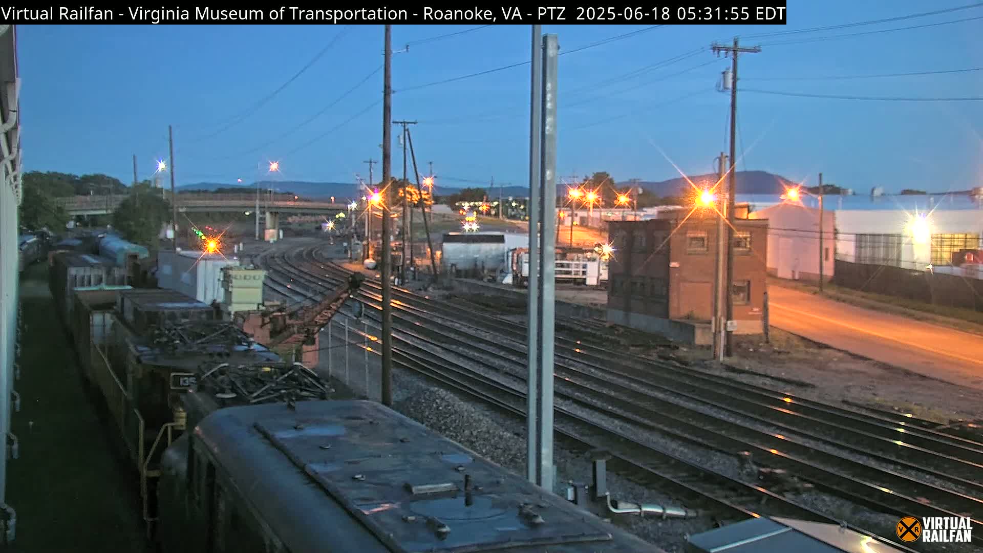 A twilight view of a train yard with multiple railroad tracks, some train cars, and industrial buildings under a mostly clear sky.