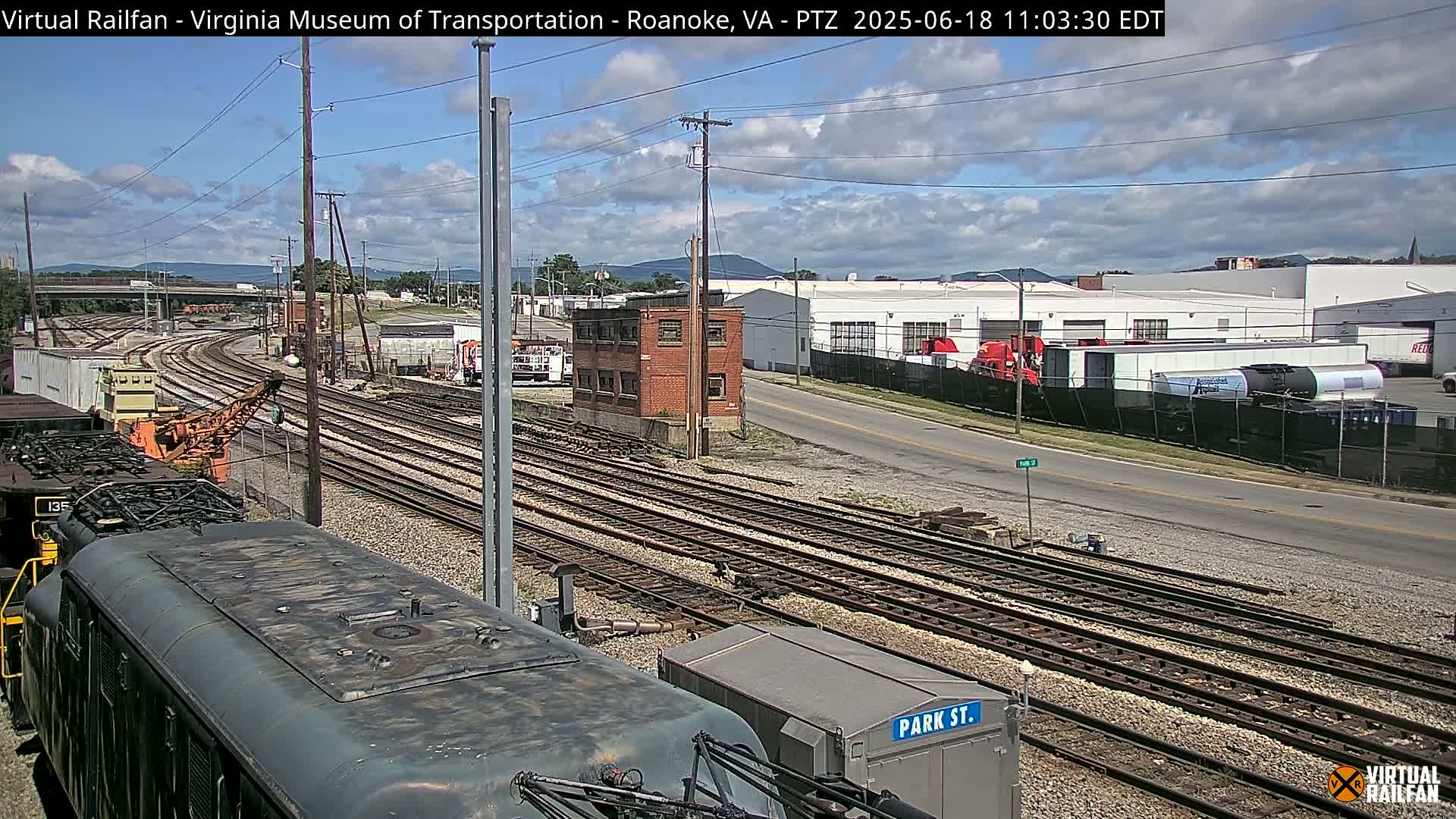 A mostly sunny day reveals a railway yard with multiple train tracks, a locomotive, industrial buildings, and a street intersecting the tracks.