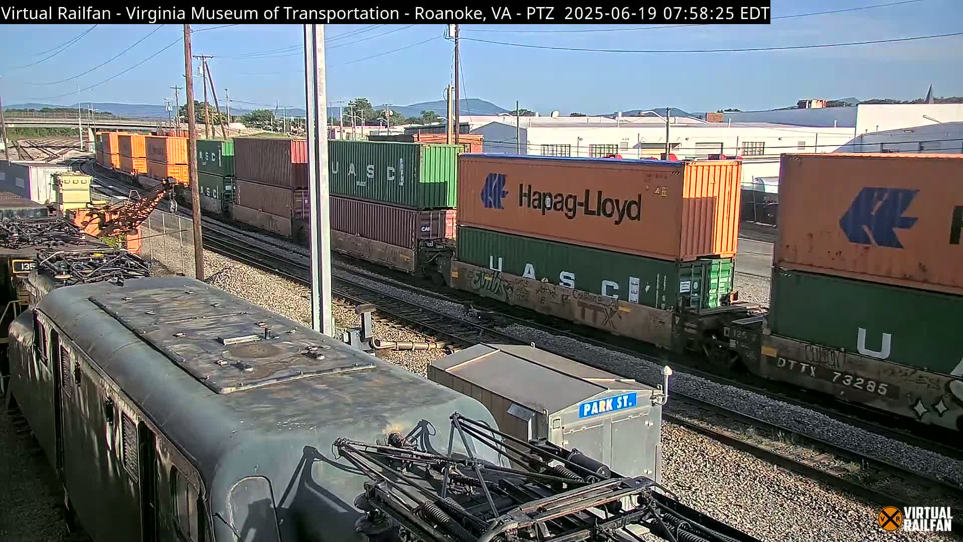A train carrying multiple shipping containers is seen on a sunny day, with another train car partially visible in the foreground.