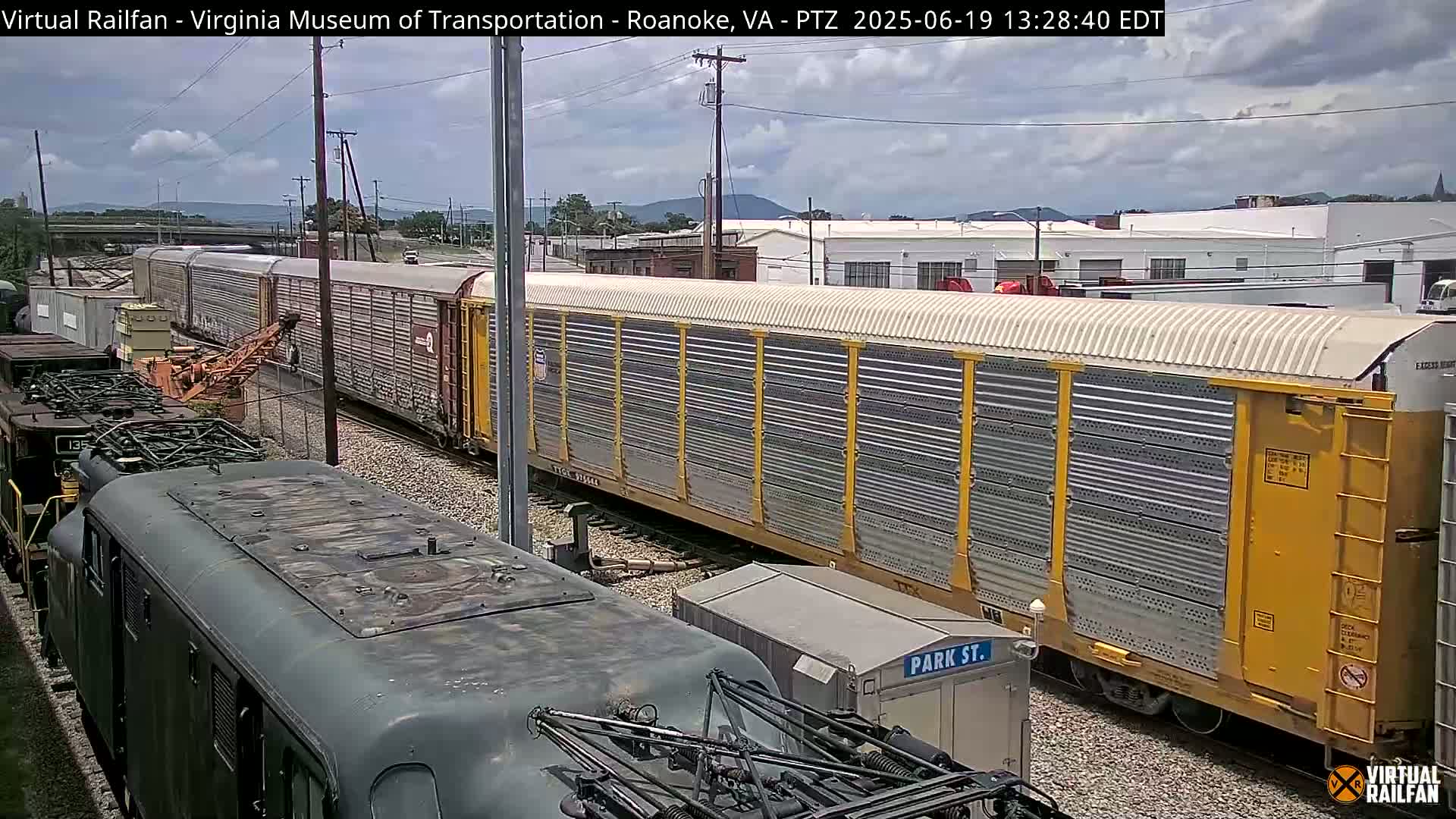 A long freight train with yellow and gray cars is partially visible, under a partly cloudy sky, next to a dark-colored locomotive.