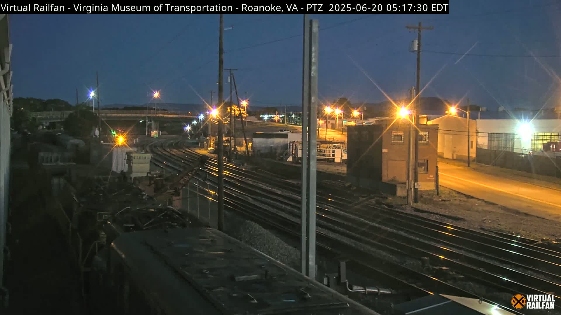 A nighttime view of a railroad yard with several sets of tracks, illuminated by streetlights and buildings, under a twilight sky.