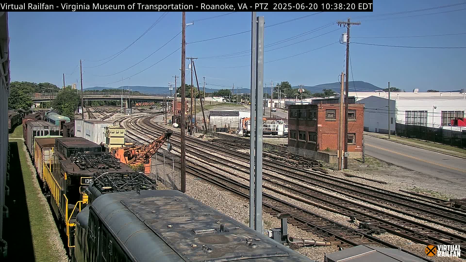 A train sits on a set of railway tracks under a clear, sunny sky, with a view of industrial buildings and mountains in the background.