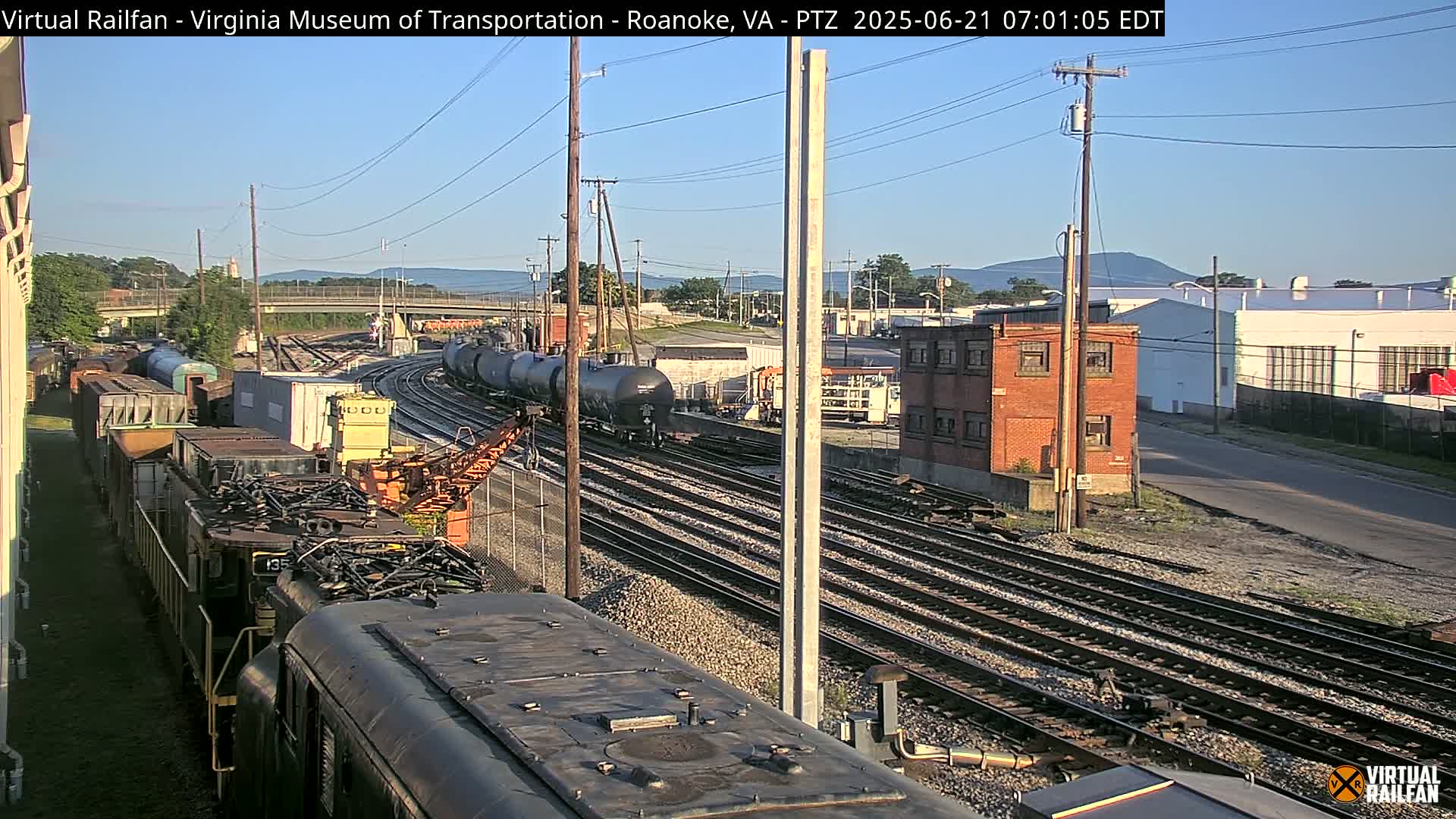 A train yard with multiple rail lines, freight cars, and locomotives under a clear, sunny sky, with distant mountains visible in the background.
