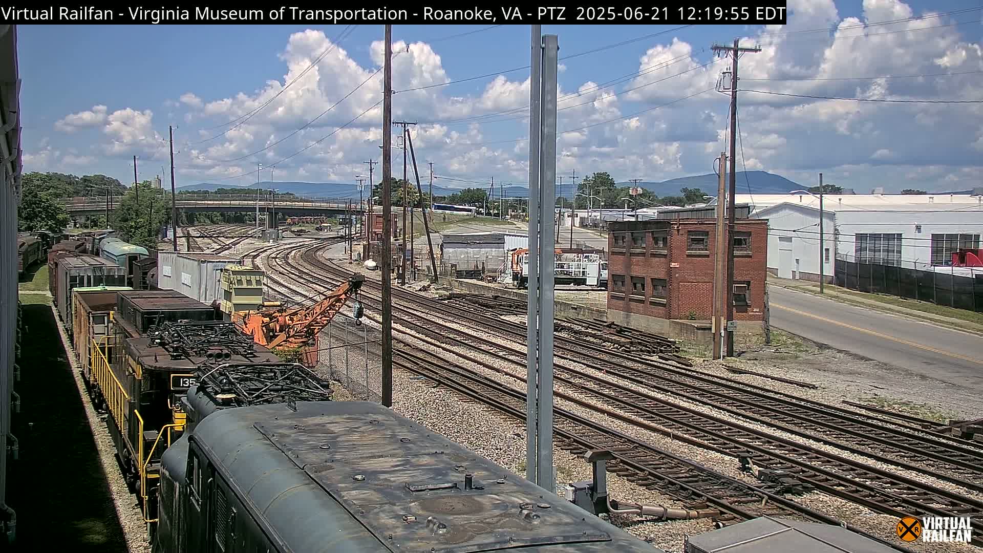 A partly cloudy day reveals a railway yard with multiple train tracks, several rail cars, and industrial buildings in the background.