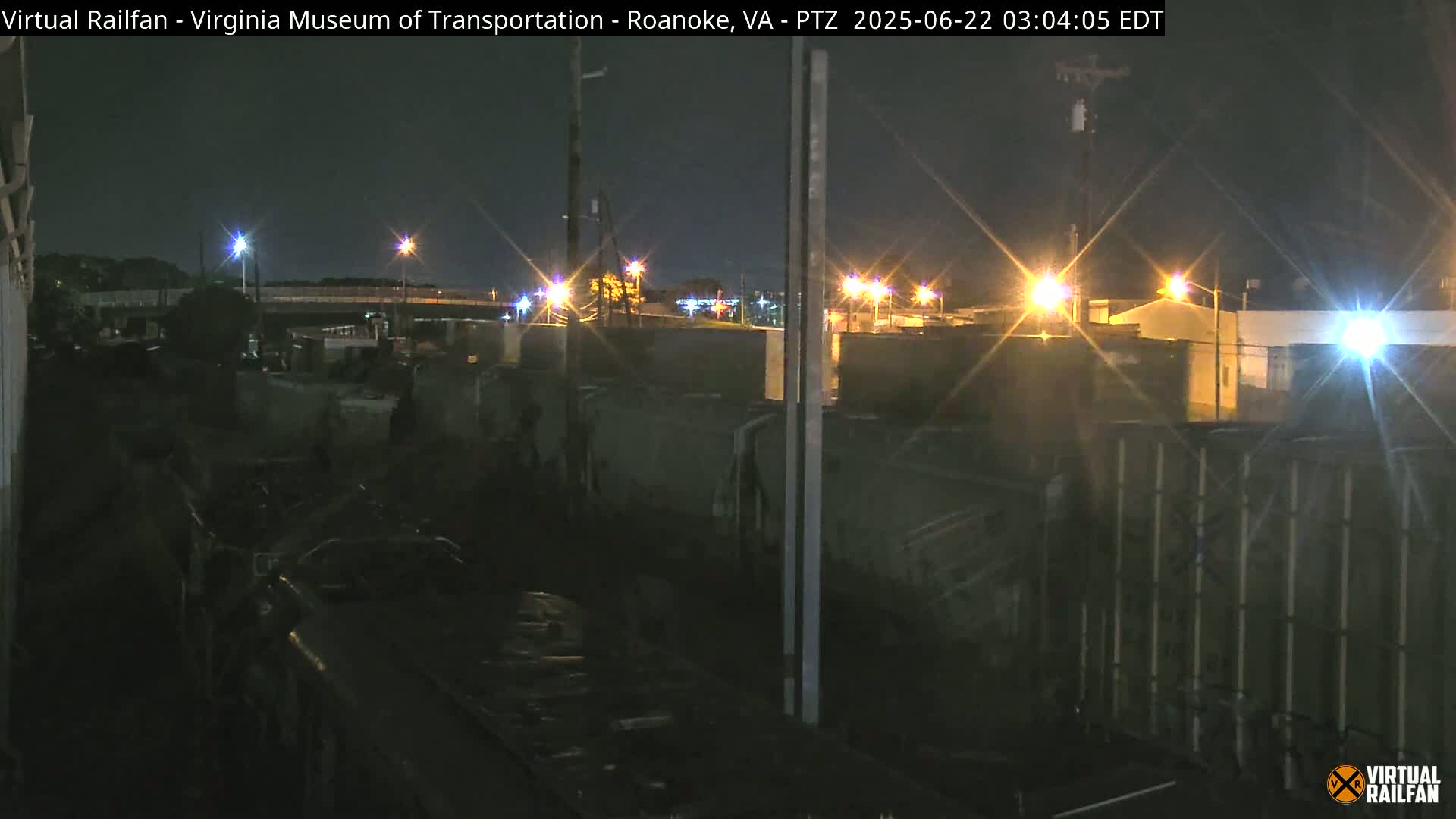 A nighttime view of a rail yard, illuminated by streetlights, with several darkened rail cars and a fence in the foreground.