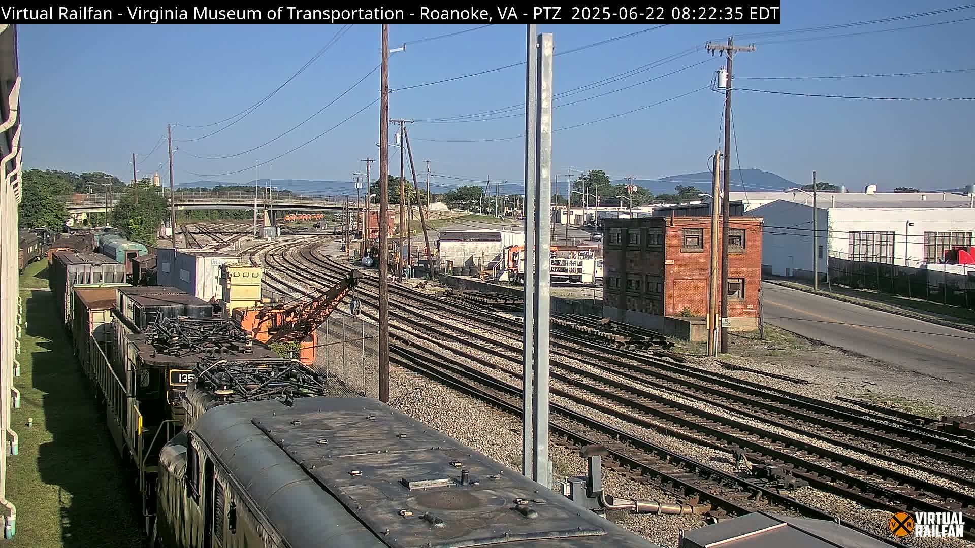 A sunny day at a train yard shows multiple sets of railroad tracks, a locomotive, and several rail cars, with distant mountains visible in the background.