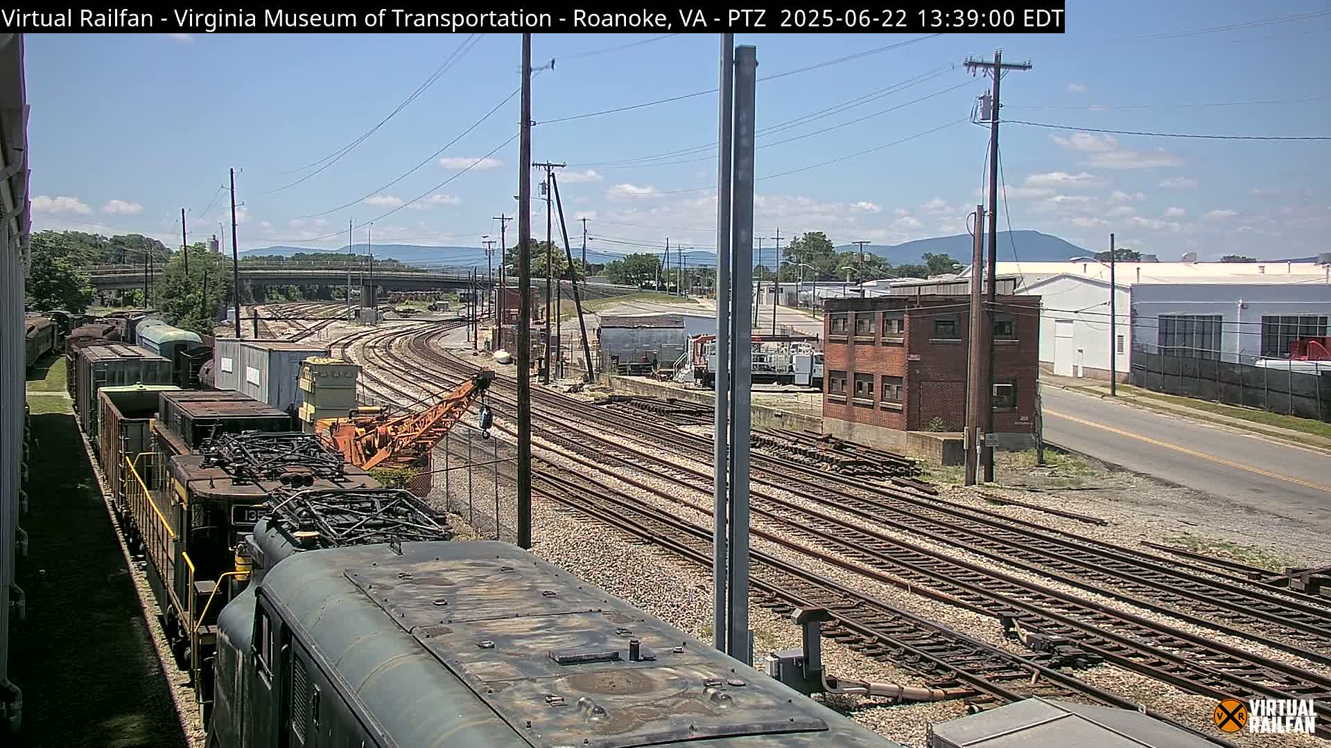 A sunny day reveals a train yard with multiple sets of tracks, a locomotive, rail cars, and industrial buildings under a mostly clear sky.