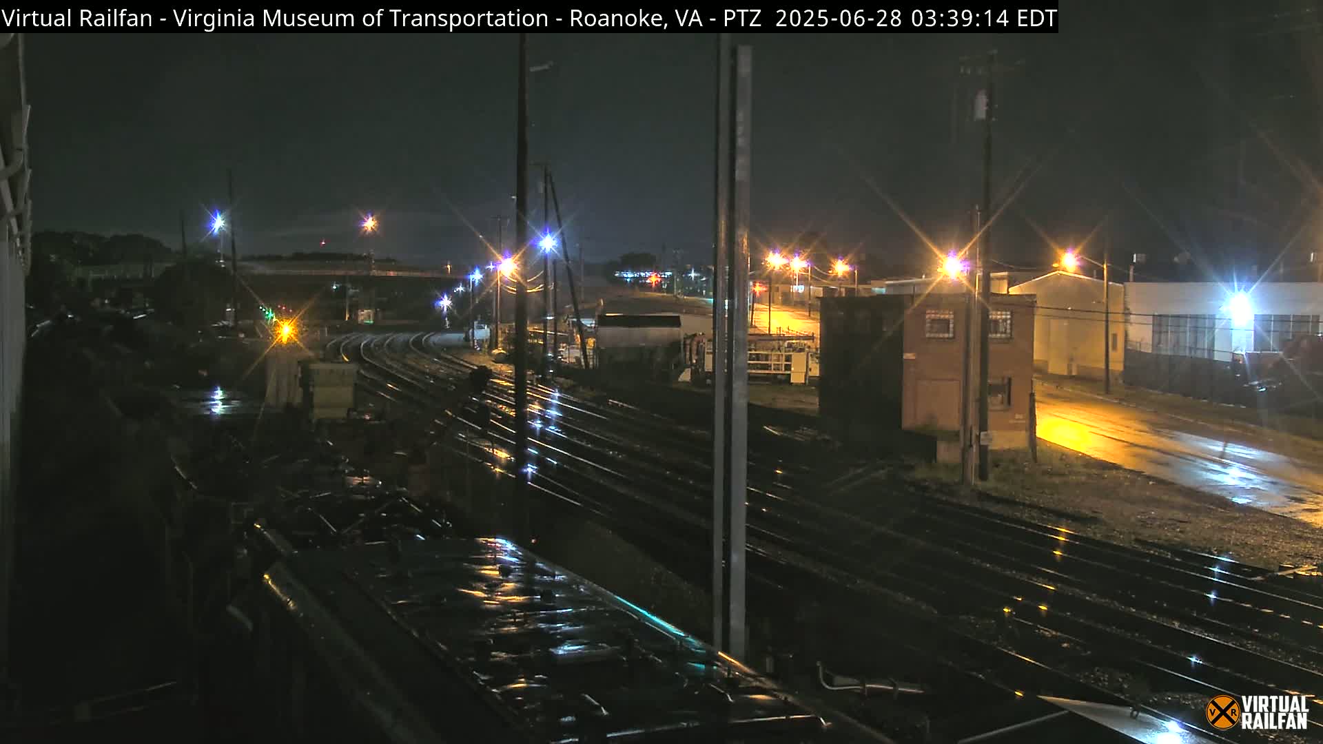 A nighttime view of rain-slicked train tracks and industrial buildings under several streetlights.