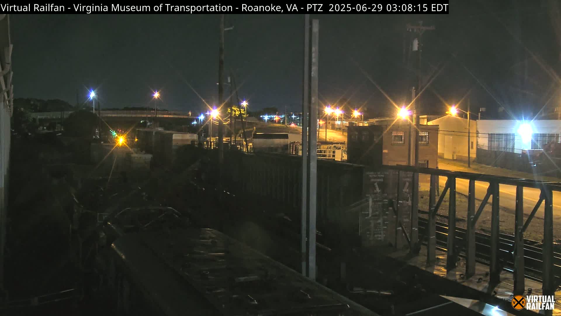 A nighttime view from a train of a railroad yard with several buildings and streetlights visible in the distance.
