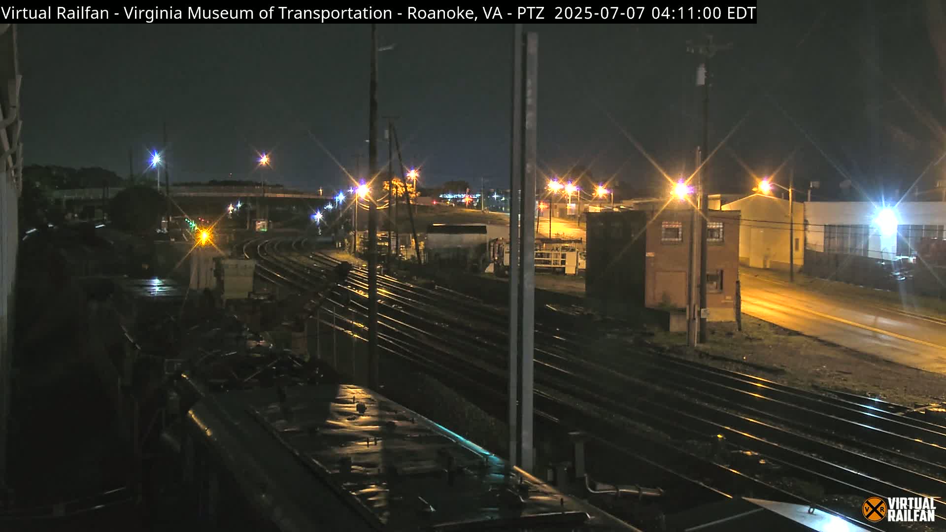 A nighttime view from atop a train car shows several sets of train tracks intersecting, with a road and buildings visible in the background under a clear sky.