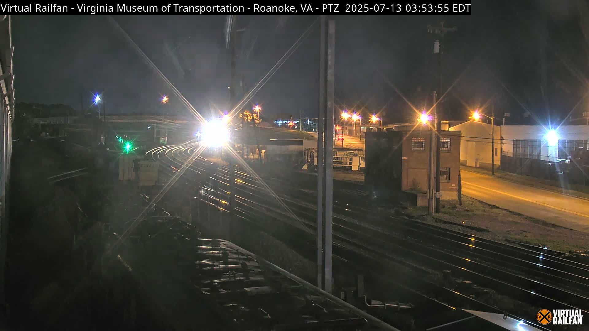 A nighttime view of a train approaching a rail yard illuminated by streetlights and train headlights.