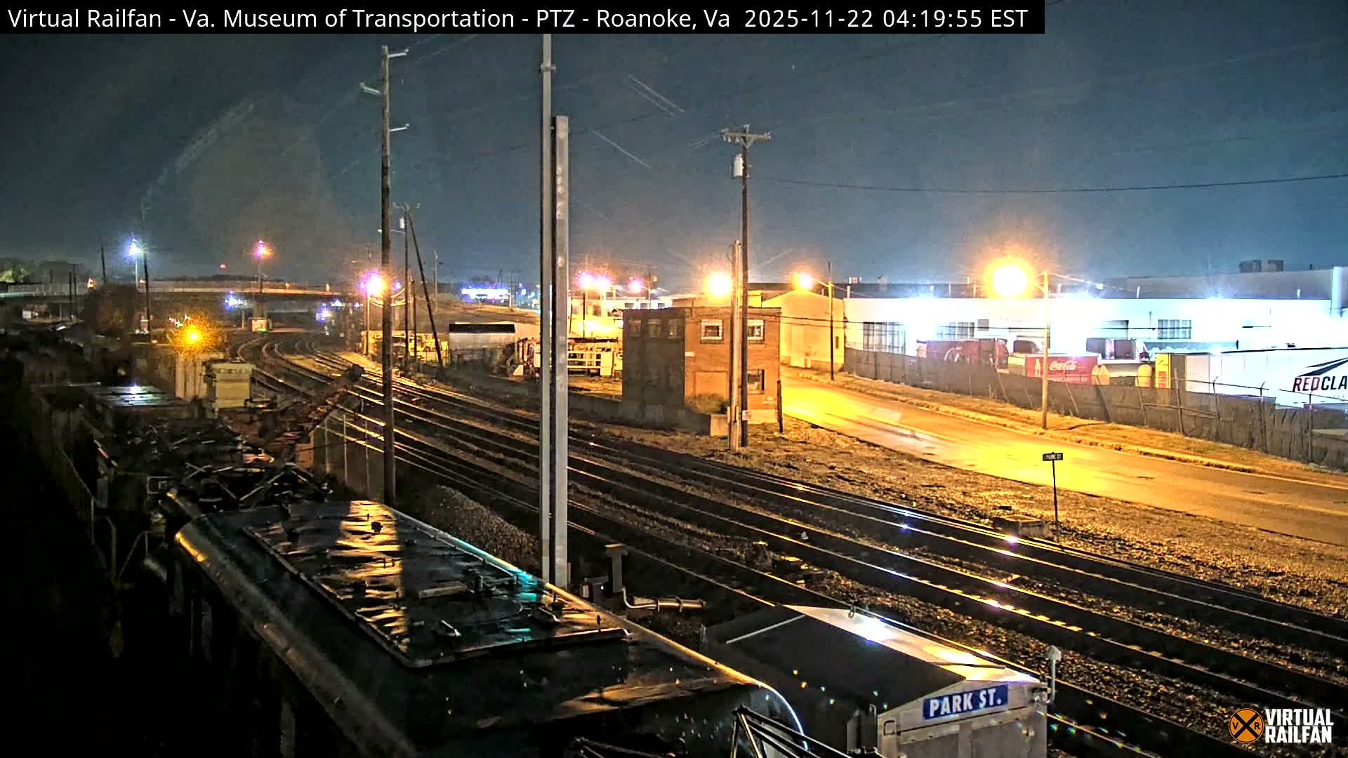 An overhead view reveals a dimly lit railway yard at night, featuring multiple tracks, various train cars, industrial buildings, and a wet road reflecting streetlights under a dark, possibly overcast sky, suggesting recent rain.
