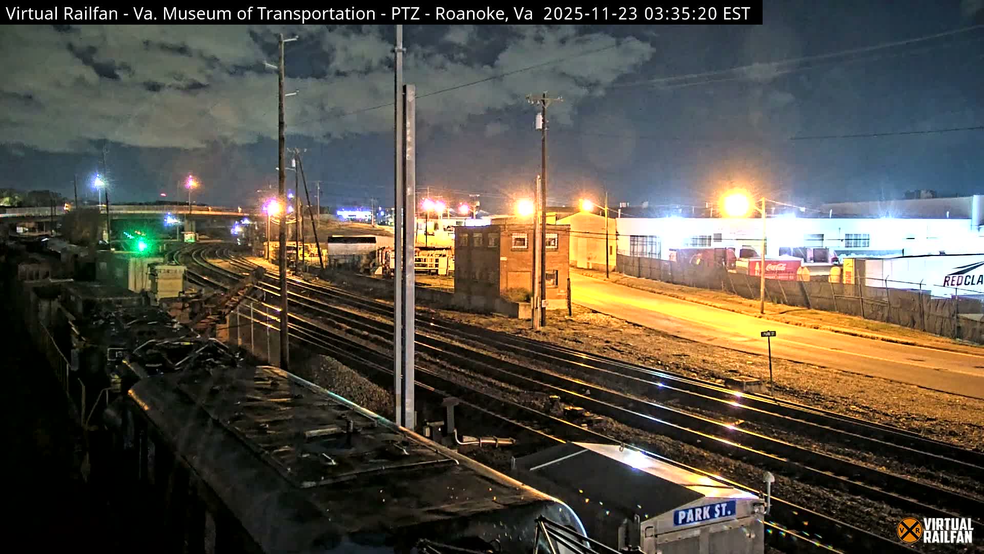 A nighttime scene reveals a sprawling railroad yard with multiple tracks and the front of a train in the foreground, illuminated by numerous streetlights and a green signal light under a partly cloudy sky, with industrial buildings and an overpass in the background.