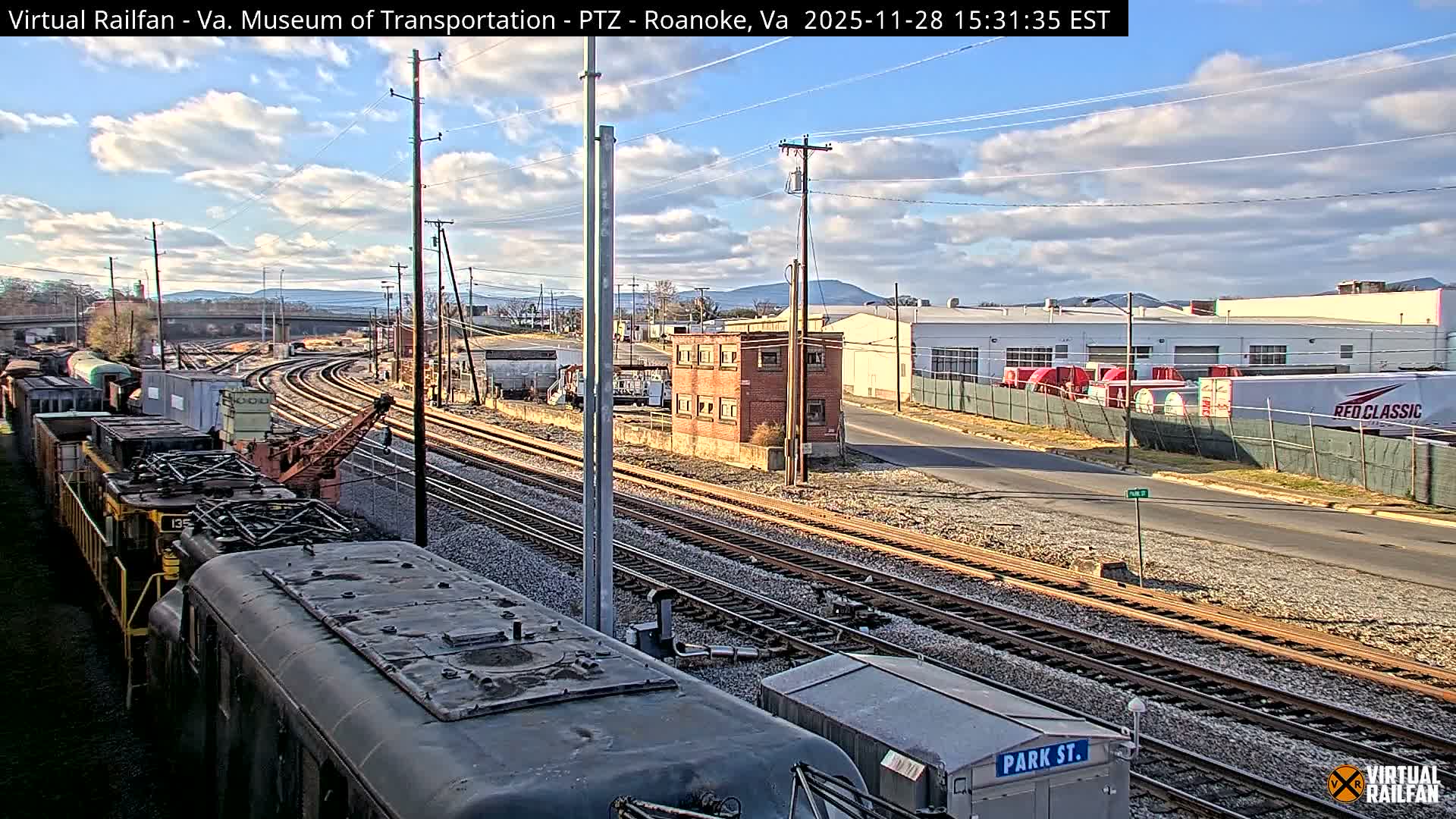 The image captures an outdoor scene of multiple train tracks curving through an industrial area with various train cars in the foreground, a small brick building, and distant mountains under a partly cloudy sky.