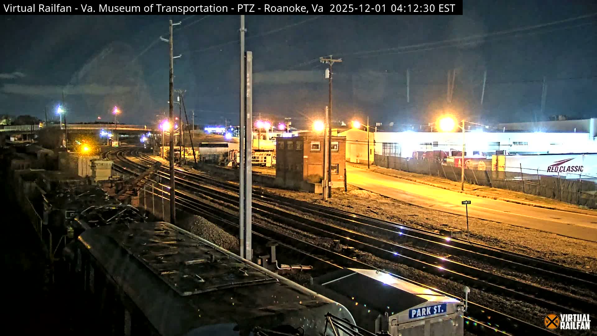 A wide, clear night view of a rail yard features multiple tracks, parked railcars in the foreground, and various illuminated buildings, all brightly lit by numerous streetlights under a dark sky.