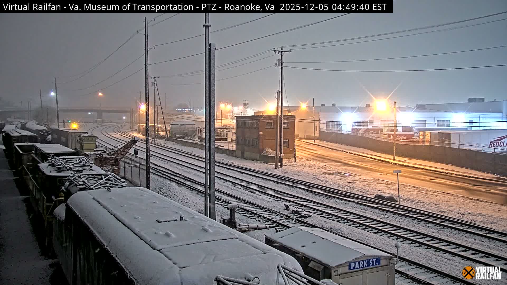 A snow-covered railway yard at night features multiple tracks, stationary trains, an old brick building, and distant industrial structures under an overcast sky.