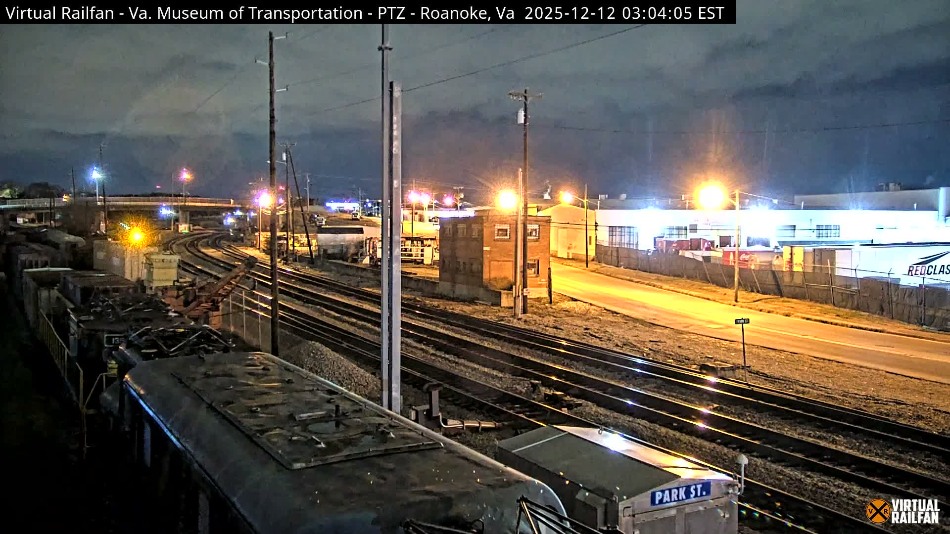 An elevated view captures a sprawling rail yard with multiple tracks and parked train cars in the foreground, illuminated by numerous artificial lights alongside a small brick building, an adjacent road, and industrial structures in the distance, all under a cloudy night sky.
