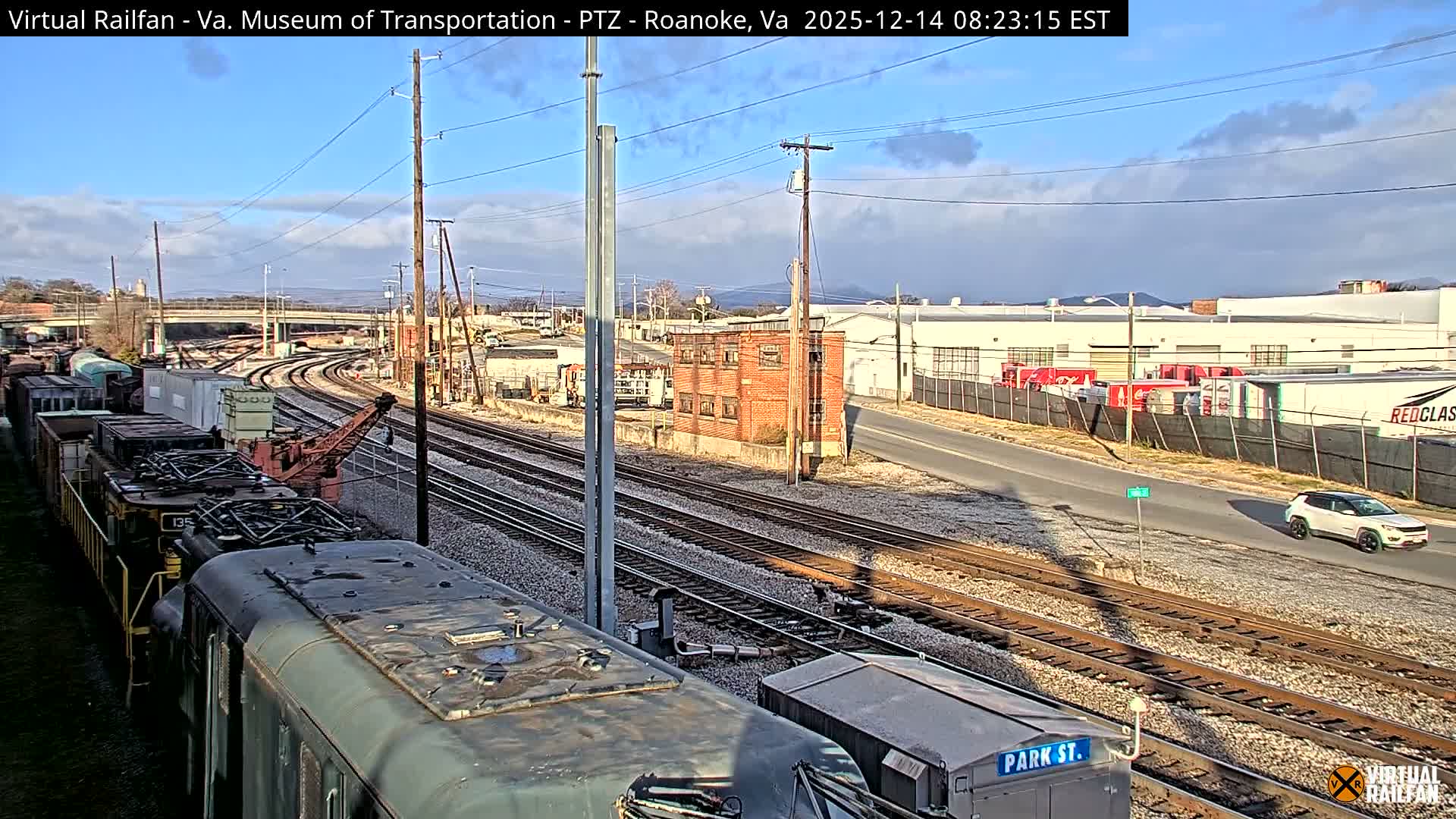 An elevated view captures a sprawling rail yard with multiple tracks and parked train cars in the foreground, illuminated by numerous artificial lights alongside a small brick building, an adjacent road, and industrial structures in the distance, all under a cloudy night sky.
