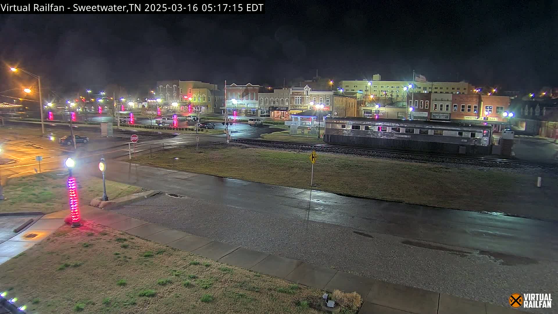 A wet, nighttime street scene shows a small town square with buildings, a train car on tracks, and decorative lights along a roadway.
