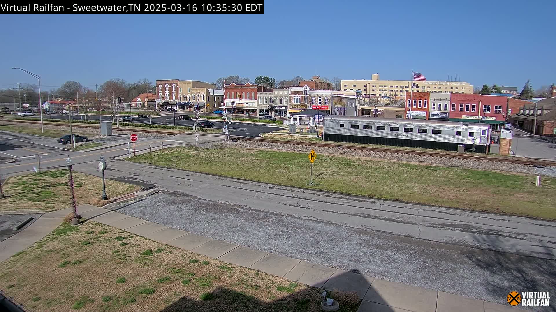 A silver passenger train sits on railway tracks in front of a town square with several brick buildings under a clear blue sky.