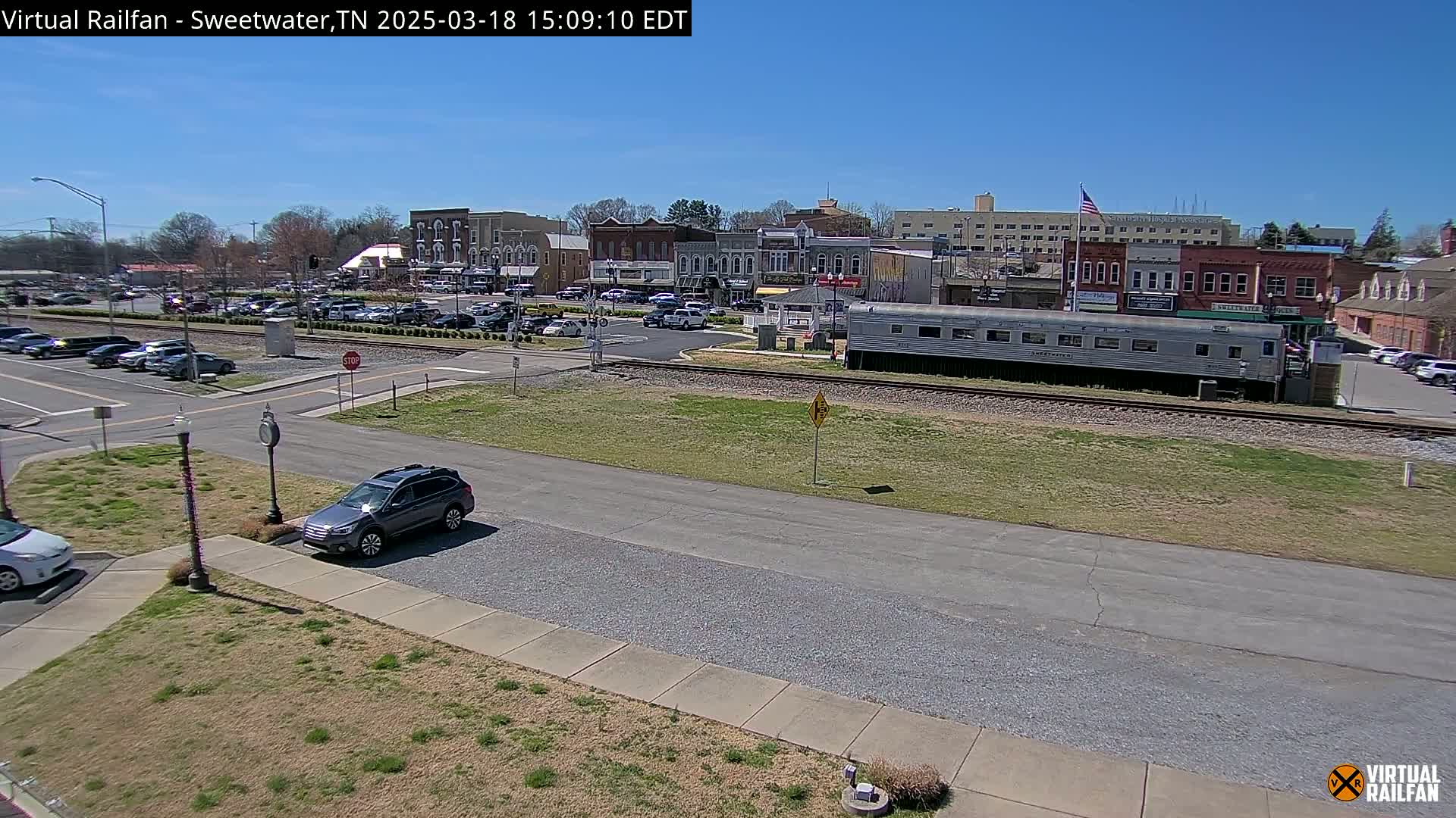 A mostly sunny day reveals a small town square with parked cars, a railroad track, and a silver passenger car; in the background, several buildings line the street.