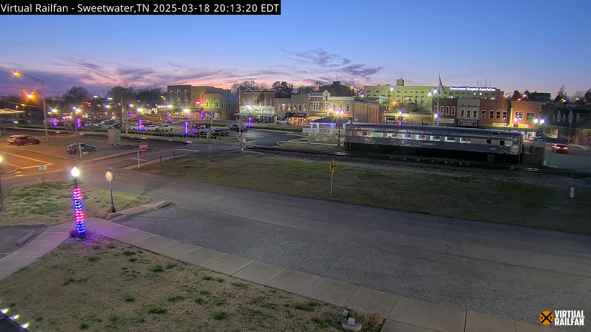 A twilight view of a town square with parked cars, streetlights, and a train car on tracks, under a mostly clear sky.