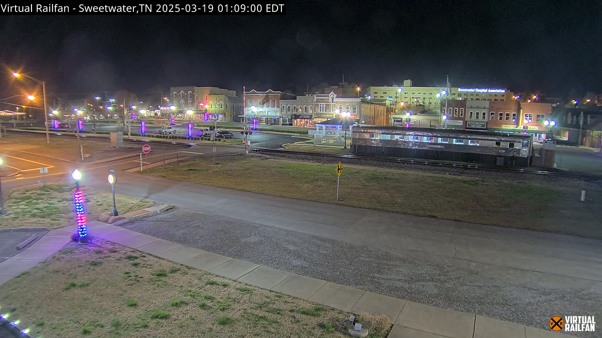 A nighttime view of a town square with several buildings, a railway car, and decorative lights illuminating the area.