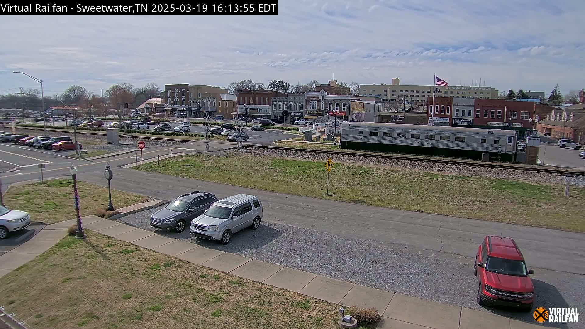 A mostly sunny view of a small town's downtown area, featuring several parked cars, a silver passenger train car, and a few brick buildings, all seen from an elevated vantage point.
