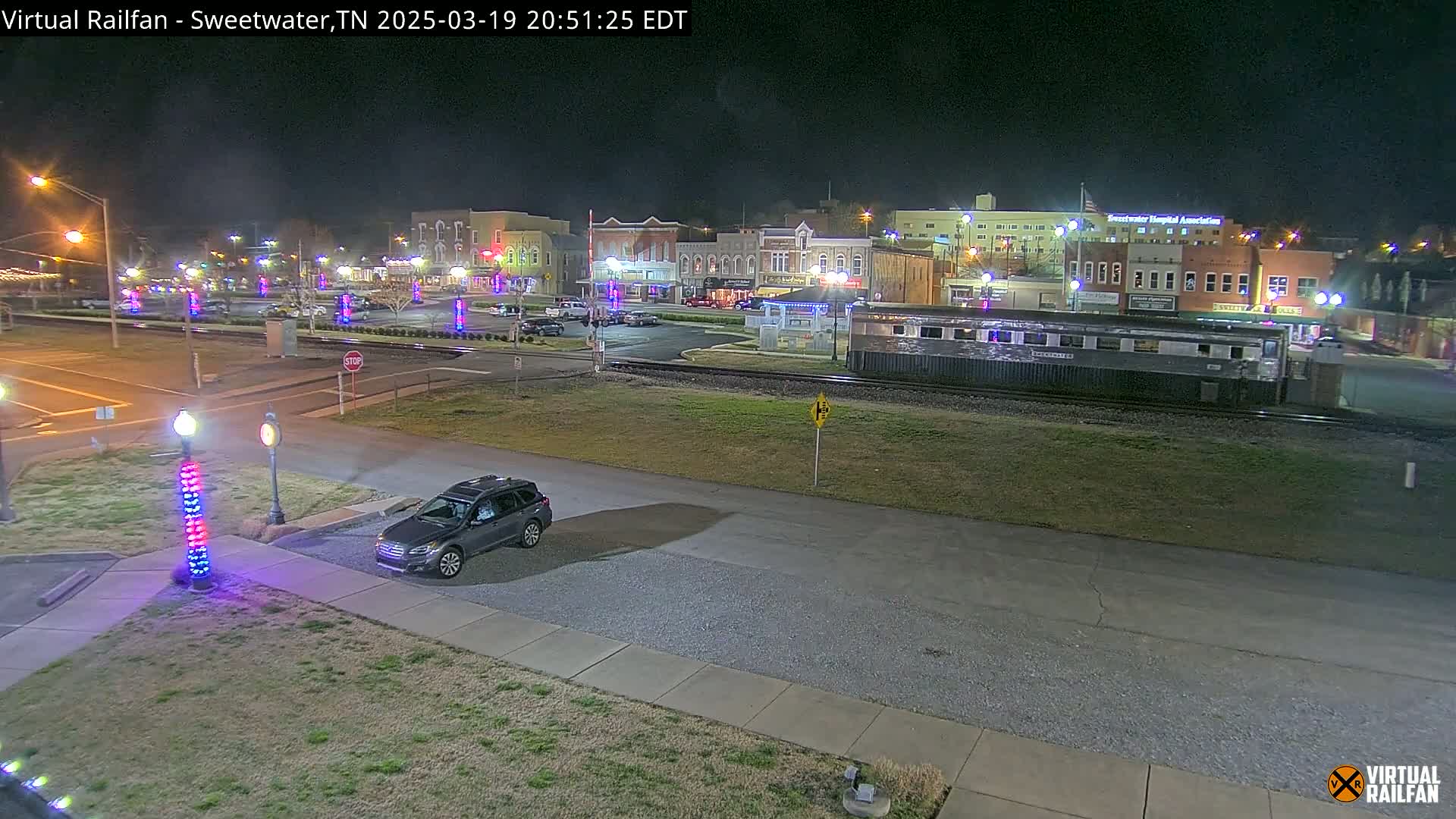 A nighttime view of a town square with several buildings, a parked SUV, and a railway car, under clear skies.
