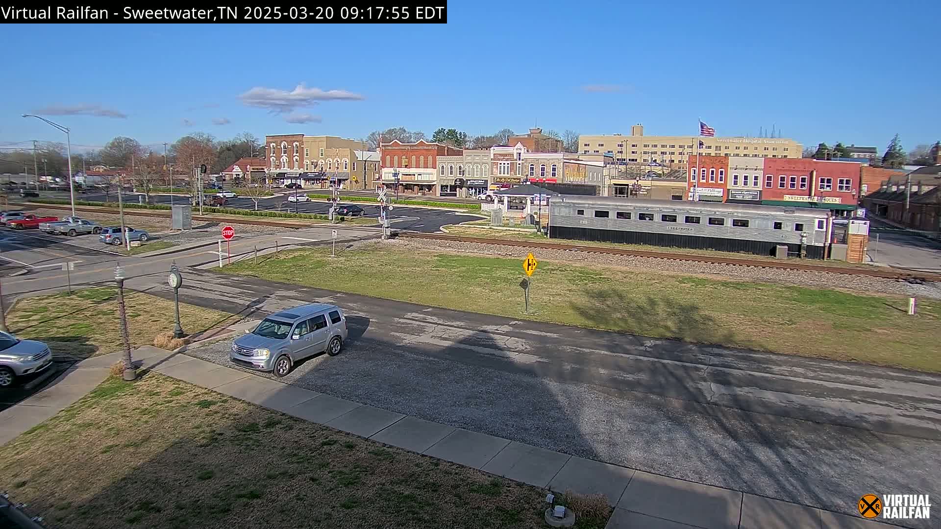 A sunny day reveals a small town's downtown area, featuring several brick buildings, a silver passenger car on train tracks, and a few cars parked in lots and on roads.