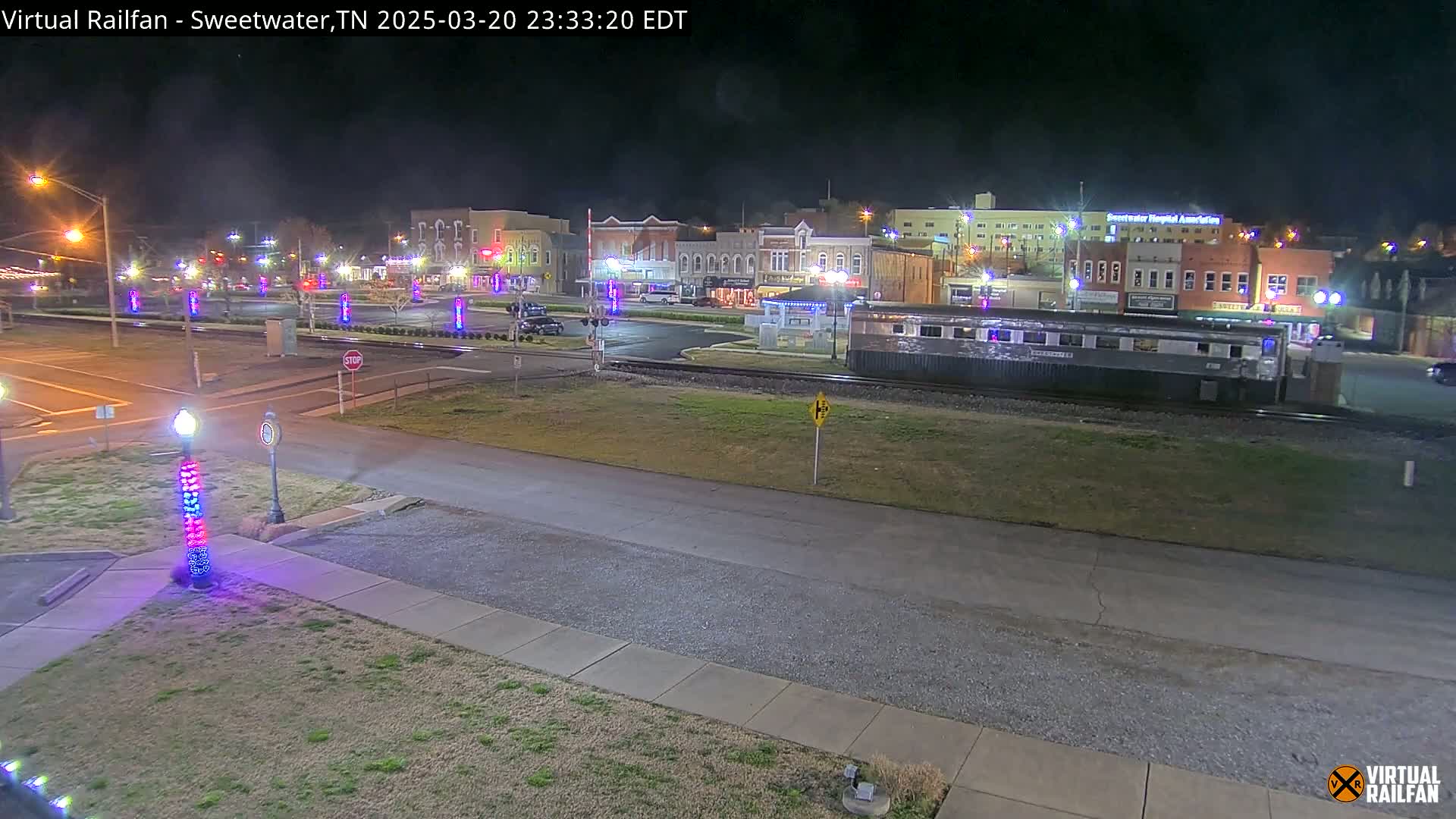 A nighttime view of a town square with a train car parked on the tracks, illuminated by streetlights and holiday decorations.