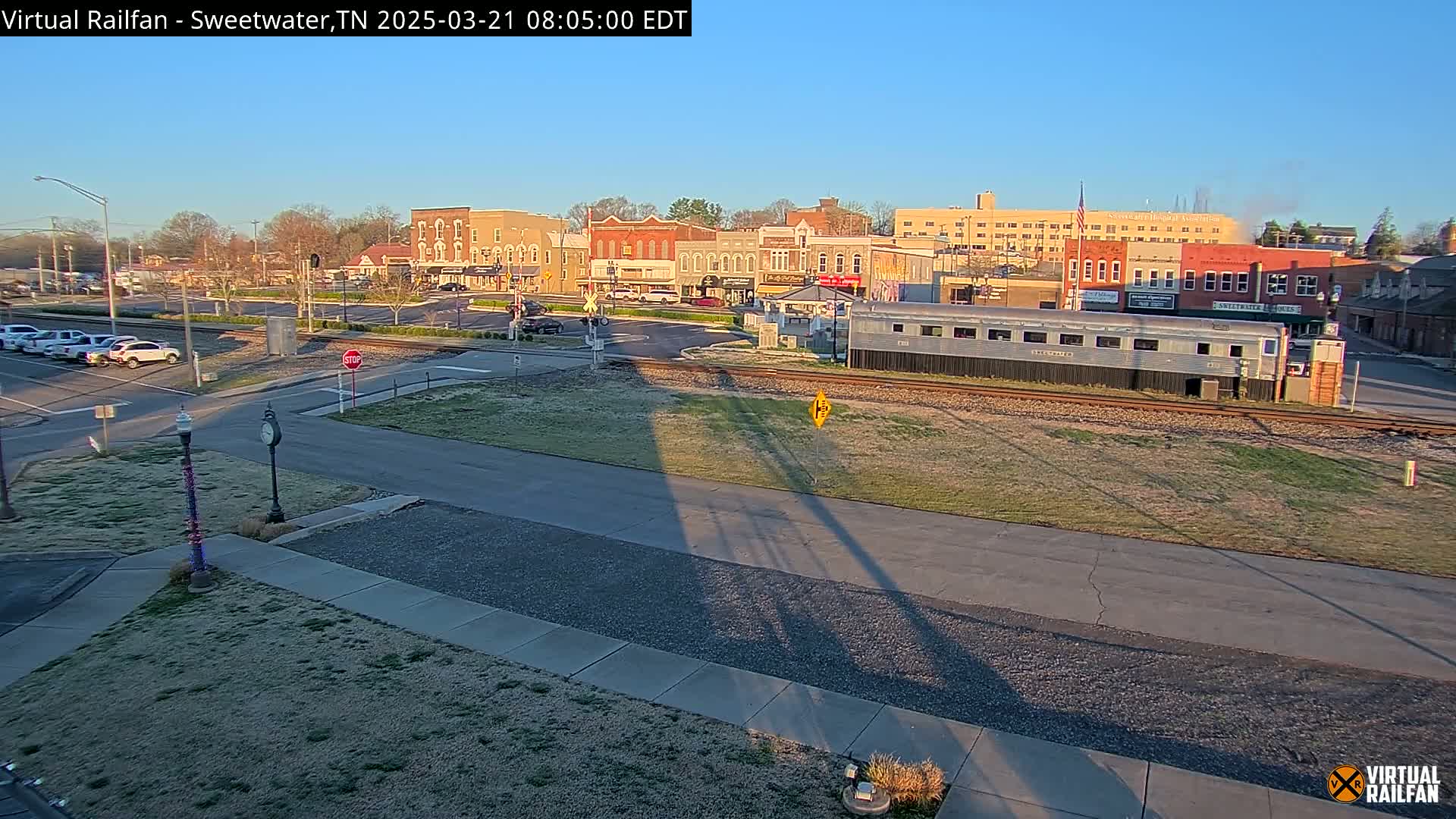 A view of a town square under a clear blue sky, with a silver railway car parked beside train tracks in the foreground.