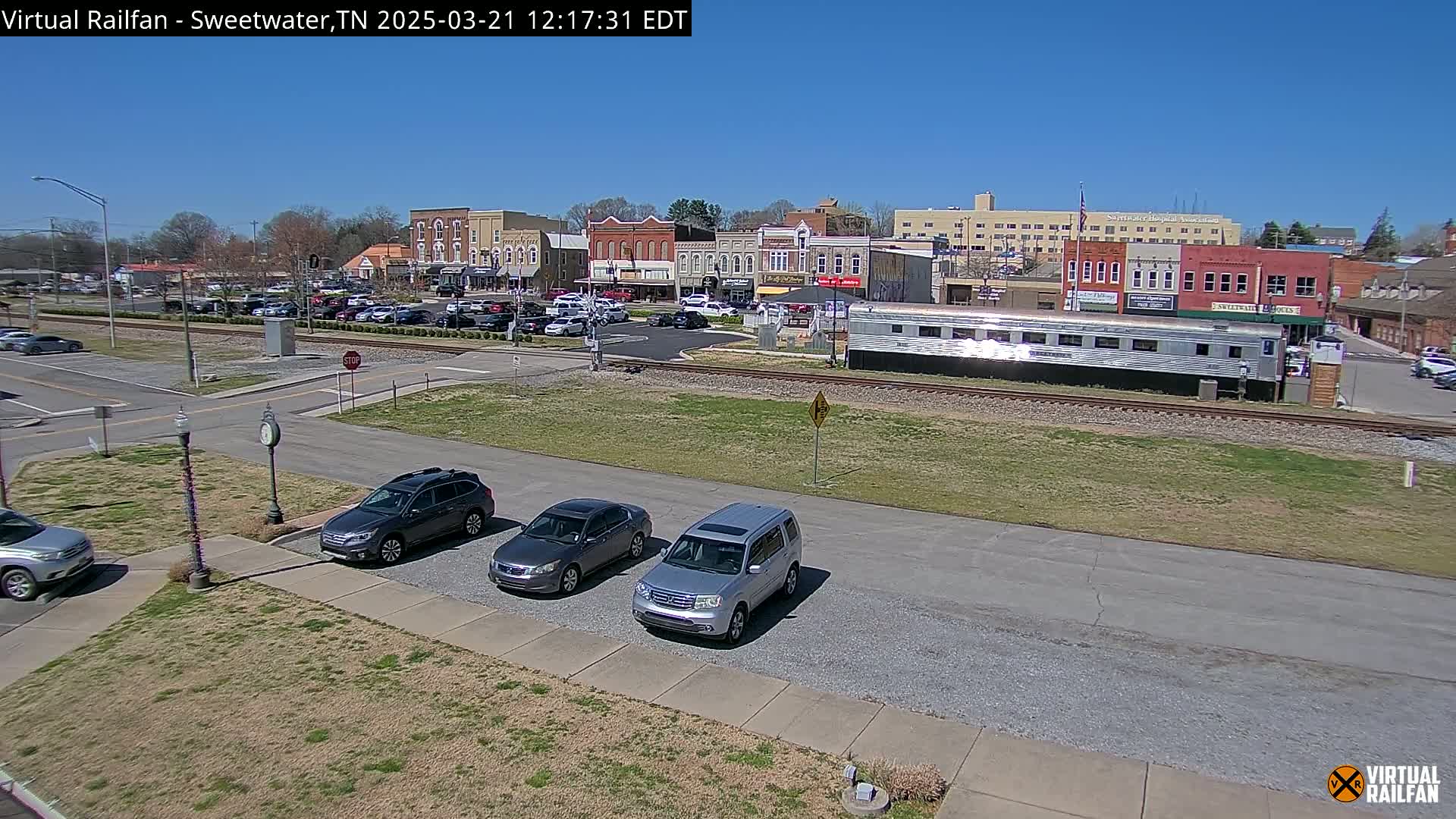 A sunny day reveals a small town's downtown area with several cars parked near a railway and a long silver train car.