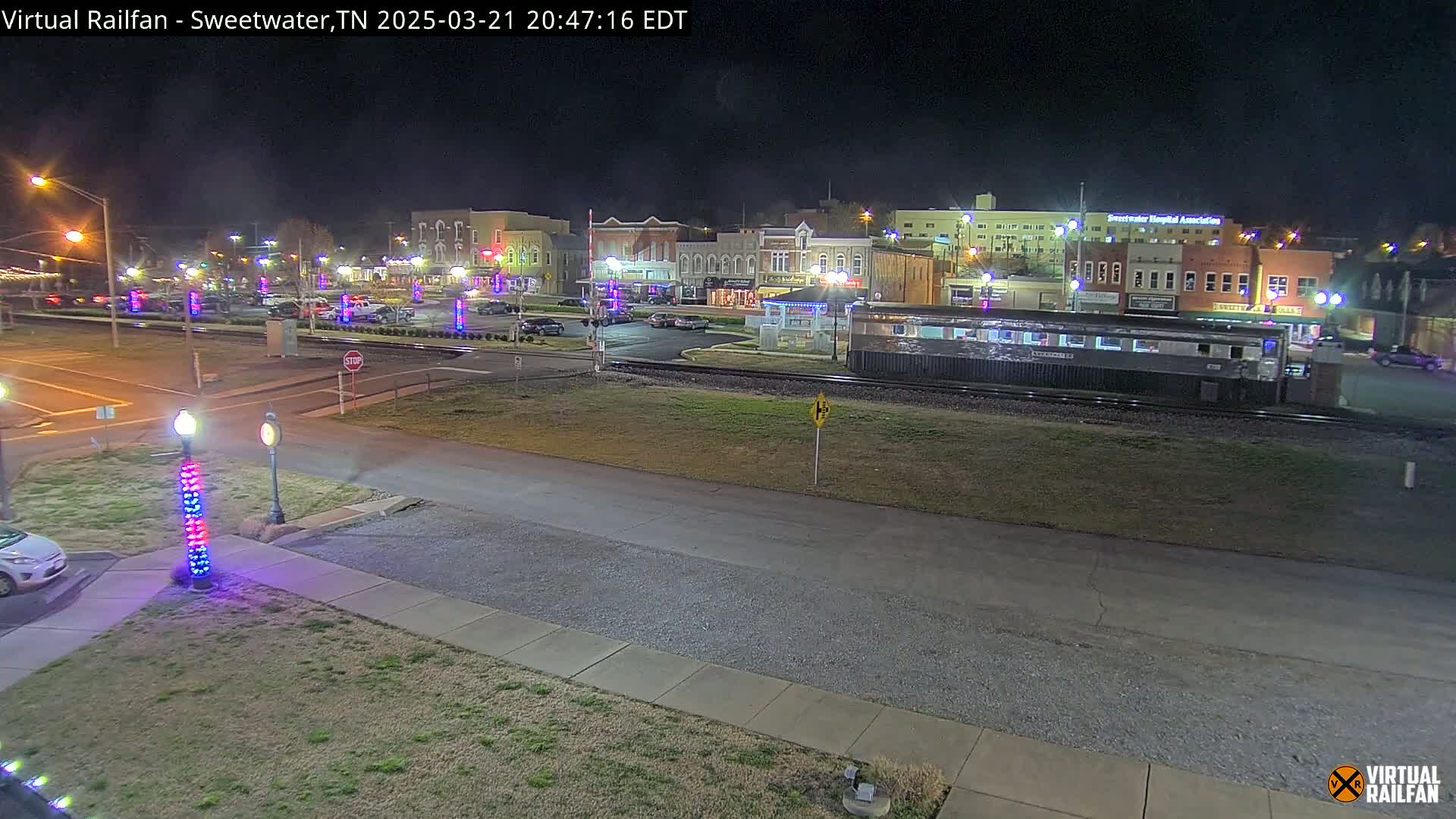 A nighttime view of a town square with several buildings, parked cars, and a train car on the tracks, under clear skies.