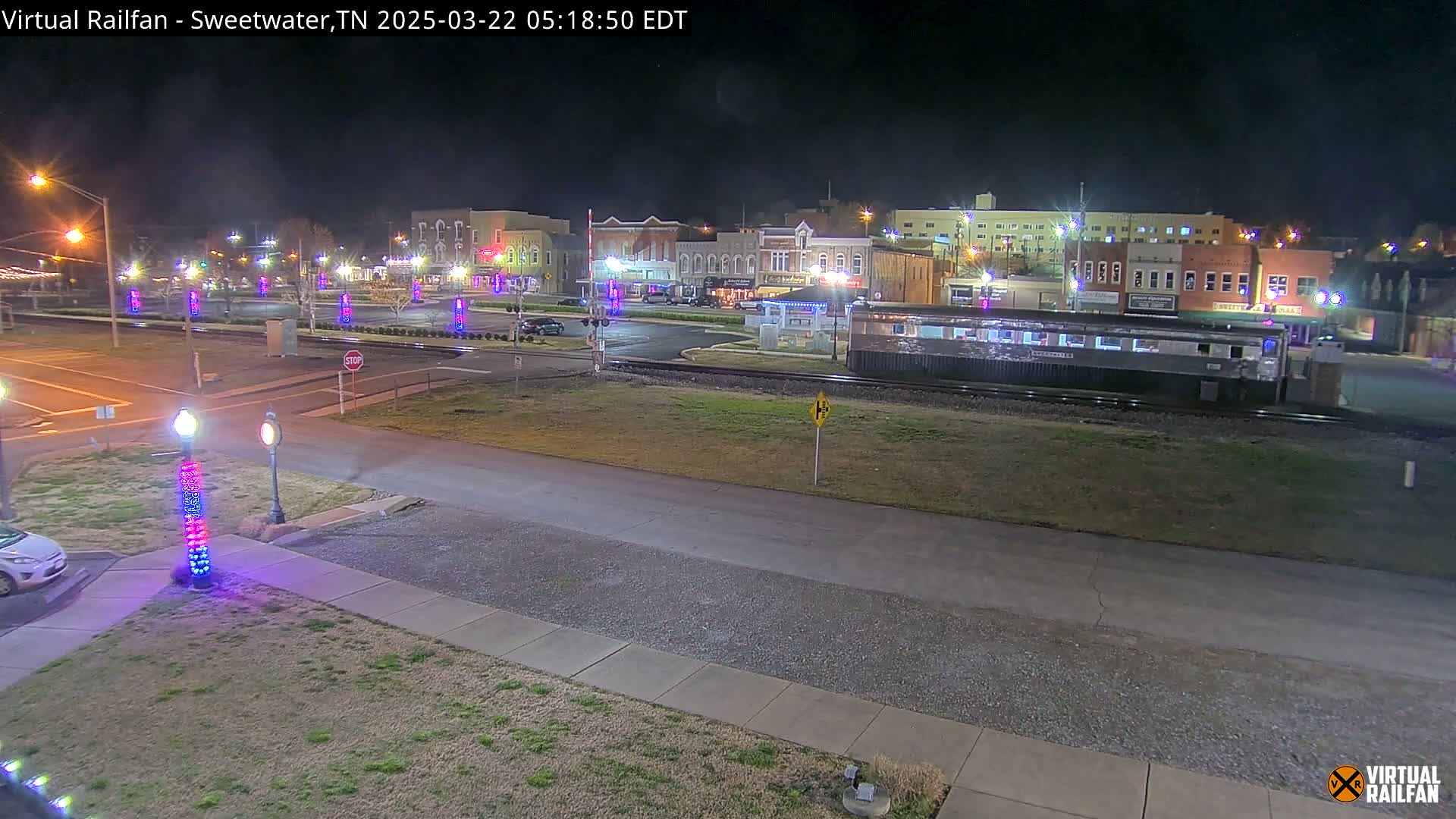 A nighttime view of a town square with several buildings, a railway track with a train car, and decorative lights illuminating the area.
