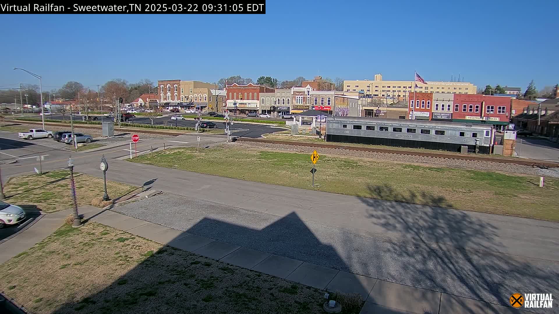 A sunny view of a small town's downtown area, featuring a long silver passenger car sitting on train tracks in front of several brick buildings.