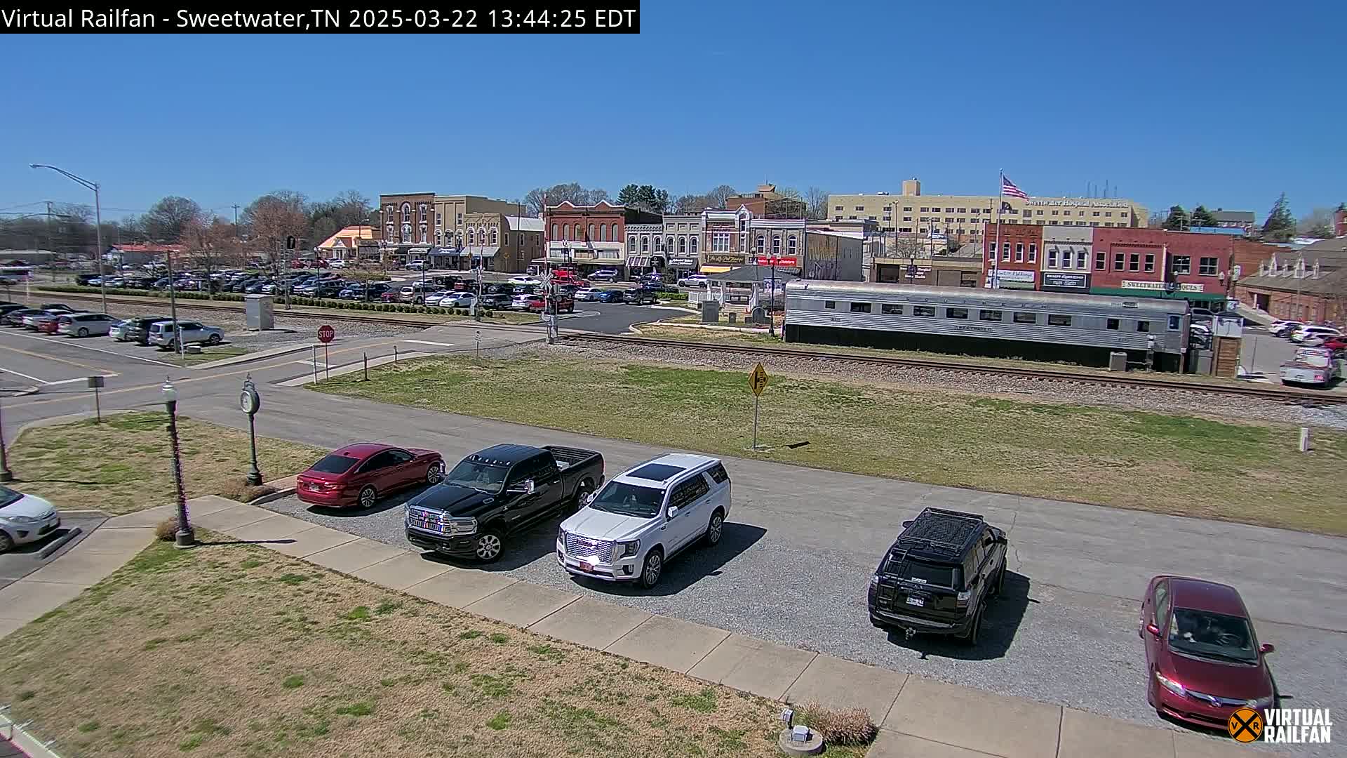 A sunny day reveals a town square with several parked cars and trucks, a train car on nearby tracks, and brick buildings lining the street.