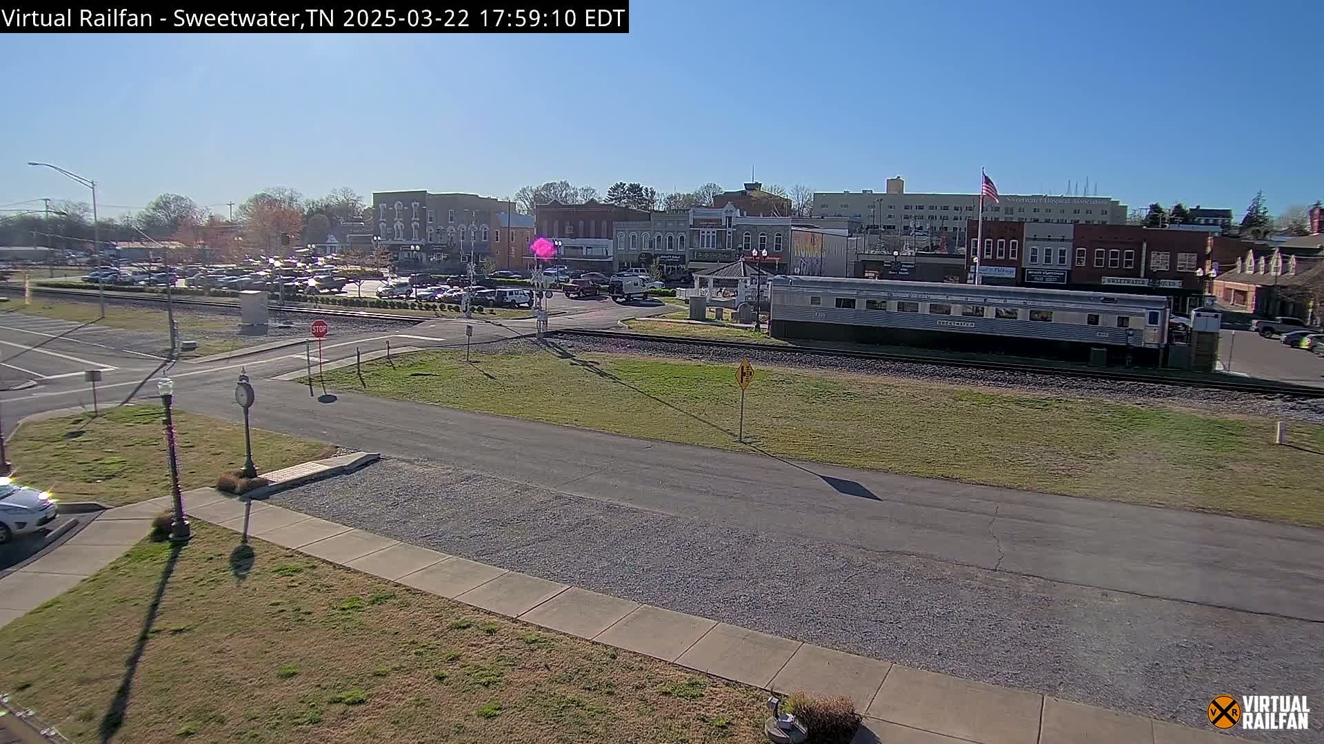A sunny day reveals a small town's square with parked cars, buildings, and a long silver train car sitting on the tracks.