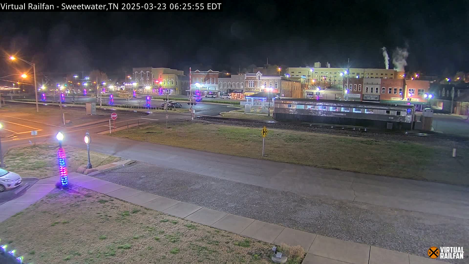 A nighttime view of a small town square with several buildings, a railway line, and a parked railcar, under clear skies.