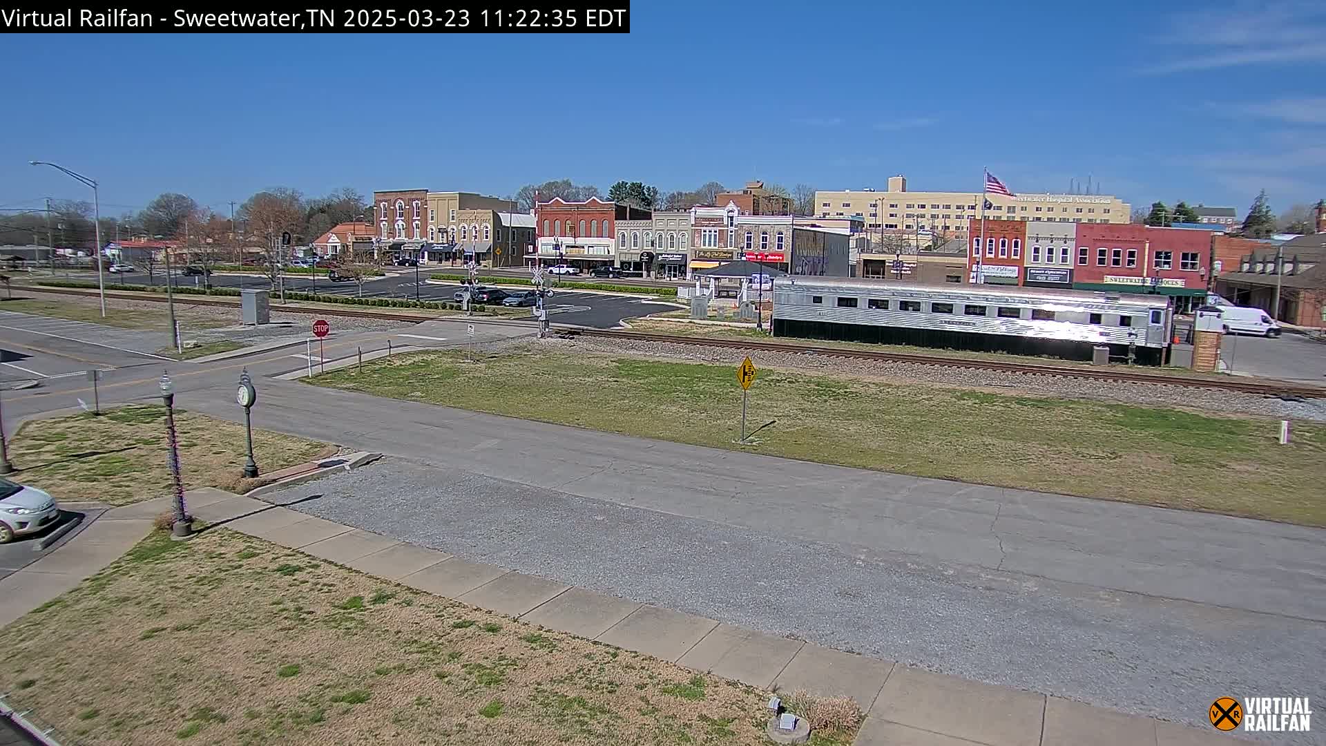 A sunny day reveals a small town's downtown area with a silver passenger rail car parked on train tracks beside a street, with cars in a parking lot and buildings lining the street.