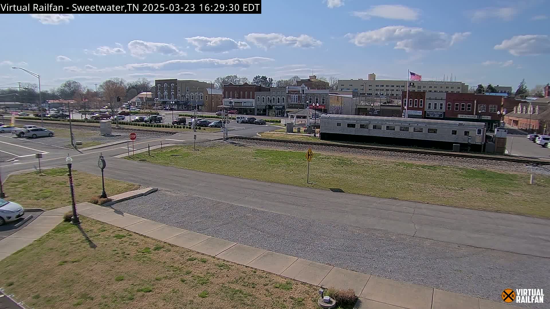 A sunny day reveals a small town's streets, buildings, and a parked passenger rail car near a railroad track.