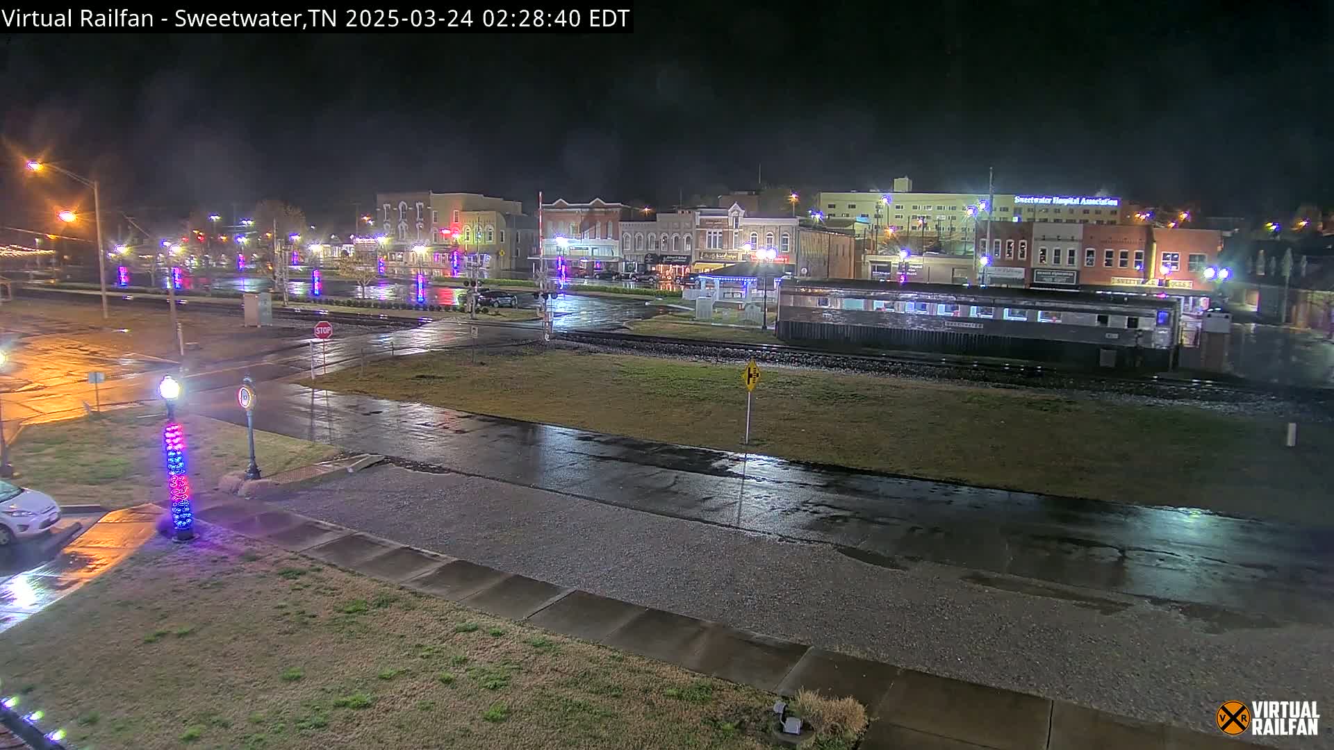 A wet, nighttime street scene shows a town square with illuminated buildings, a railway car, and decorative lights along the road.