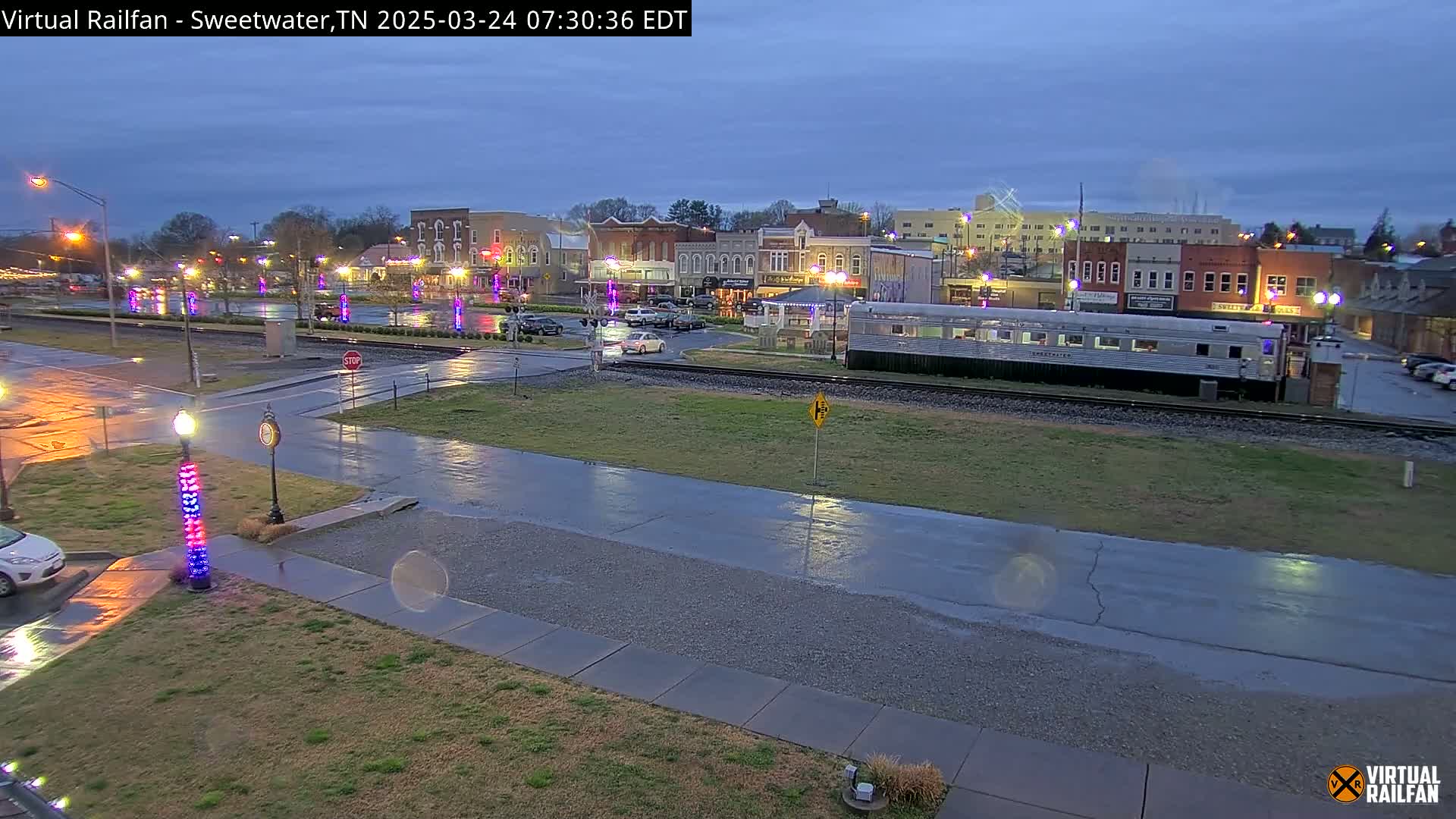 A wet, nighttime view of a town square with a passenger train car parked on the tracks in the foreground, and several buildings and vehicles visible beyond.