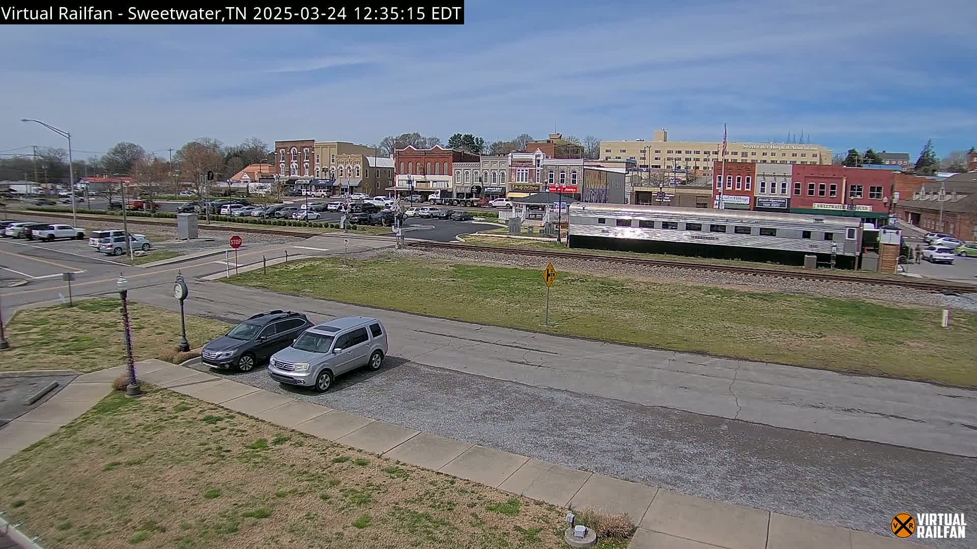 A sunny day reveals a small town's downtown area, with parked cars, a train car on tracks, and several brick buildings visible in the background.