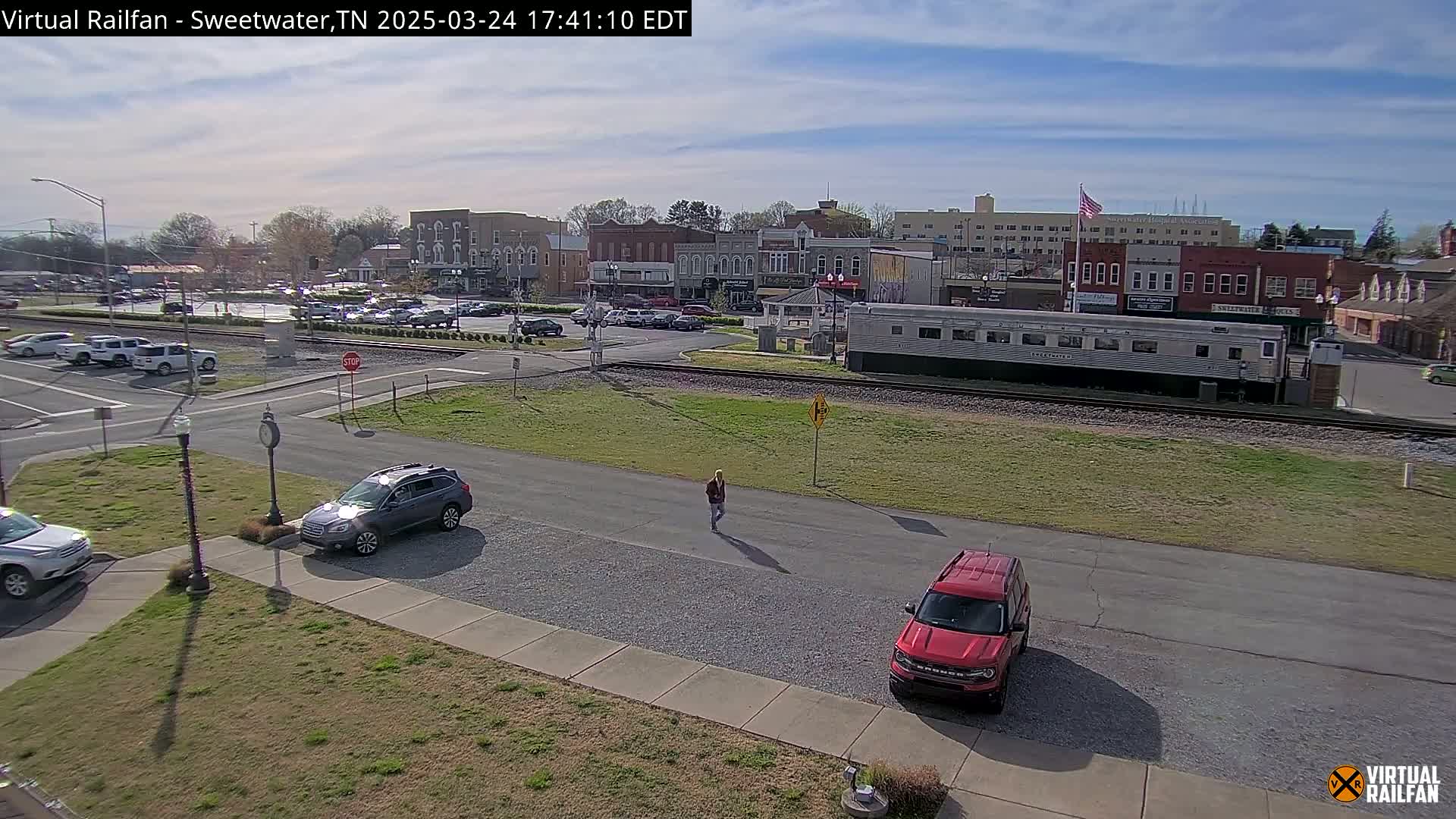 A sunny day reveals a small town square with parked cars, a pedestrian, and a long silver train car near railroad tracks.