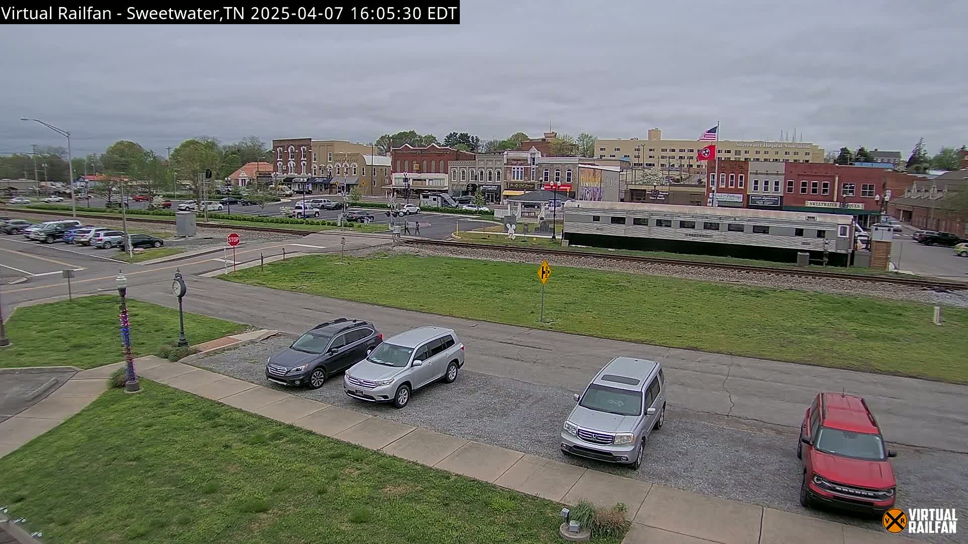 A small town square with several cars parked in a lot, a train car on the tracks, and buildings in the background under an overcast sky.