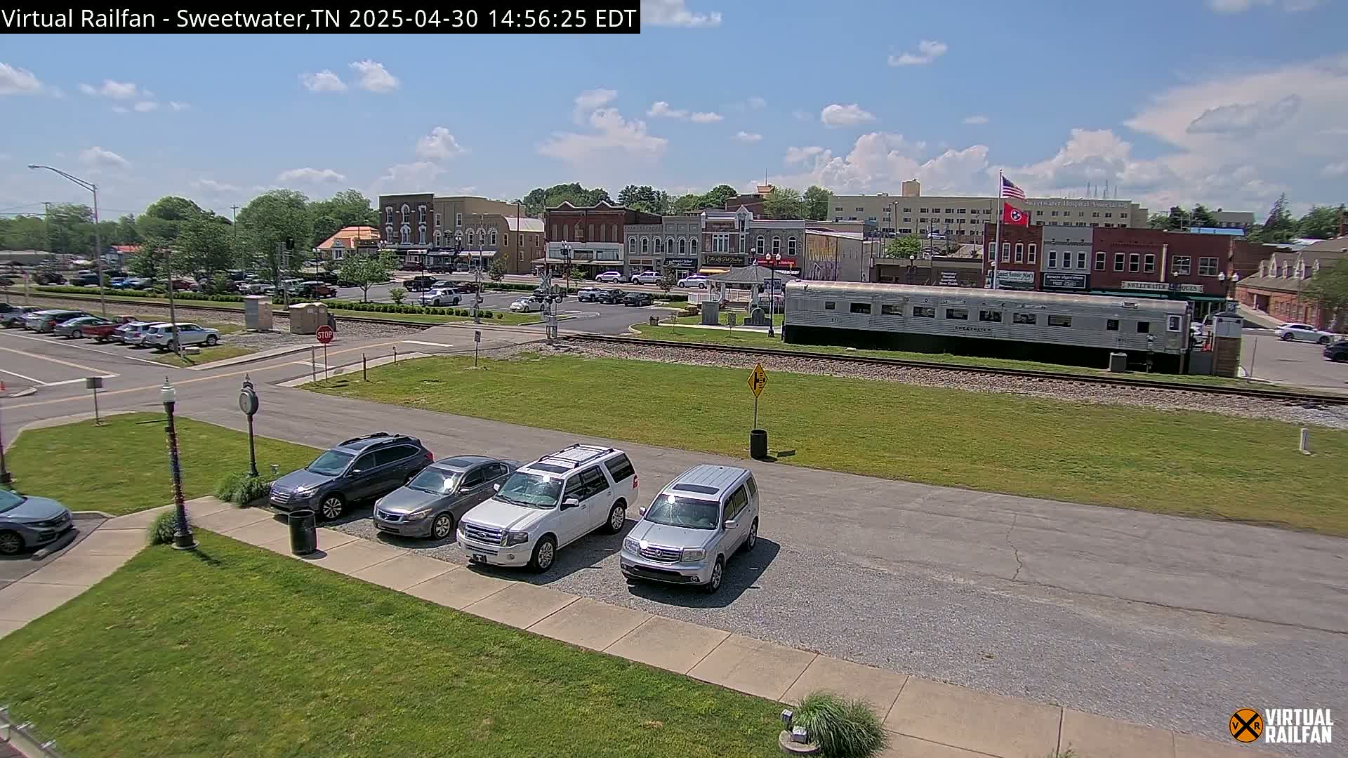 A sunny day reveals a town square with several cars parked nearby, a long railway car sitting on the tracks, and brick buildings in the background.