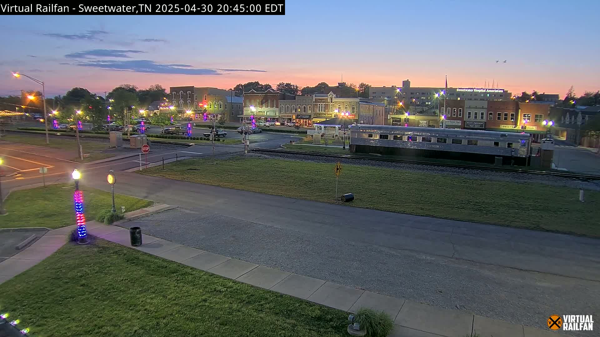 A twilight view of a town square with a silver passenger train car parked on the tracks in the foreground, under a mostly clear sky.
